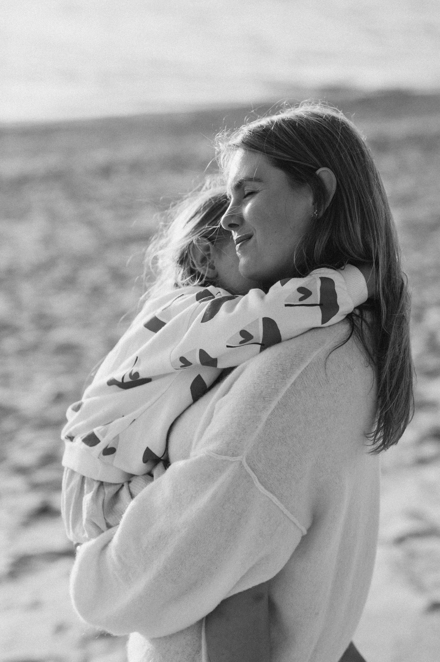 Black and white photo of a smiling woman holding a child wrapped in a patterned blanket on a beach.