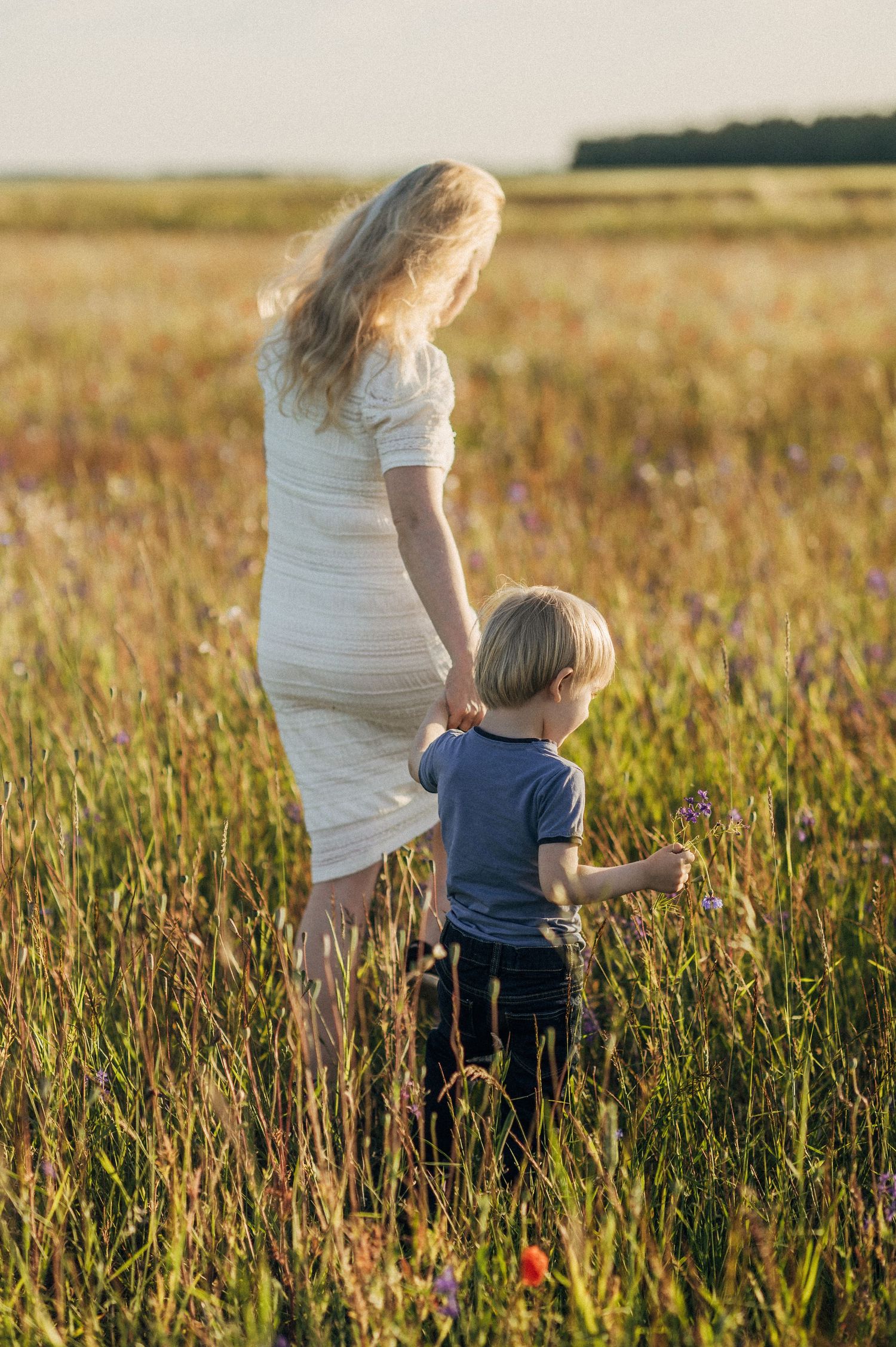 Mother and child walking through tall grass in a meadow at sunset.