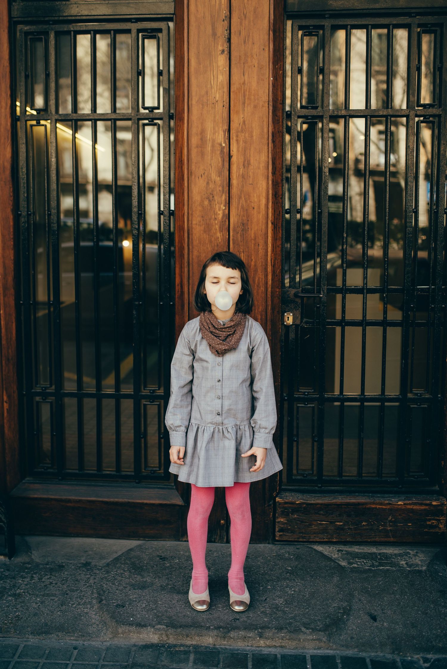 Child in grey dress and pink tights standing in front of dark wooden doors.