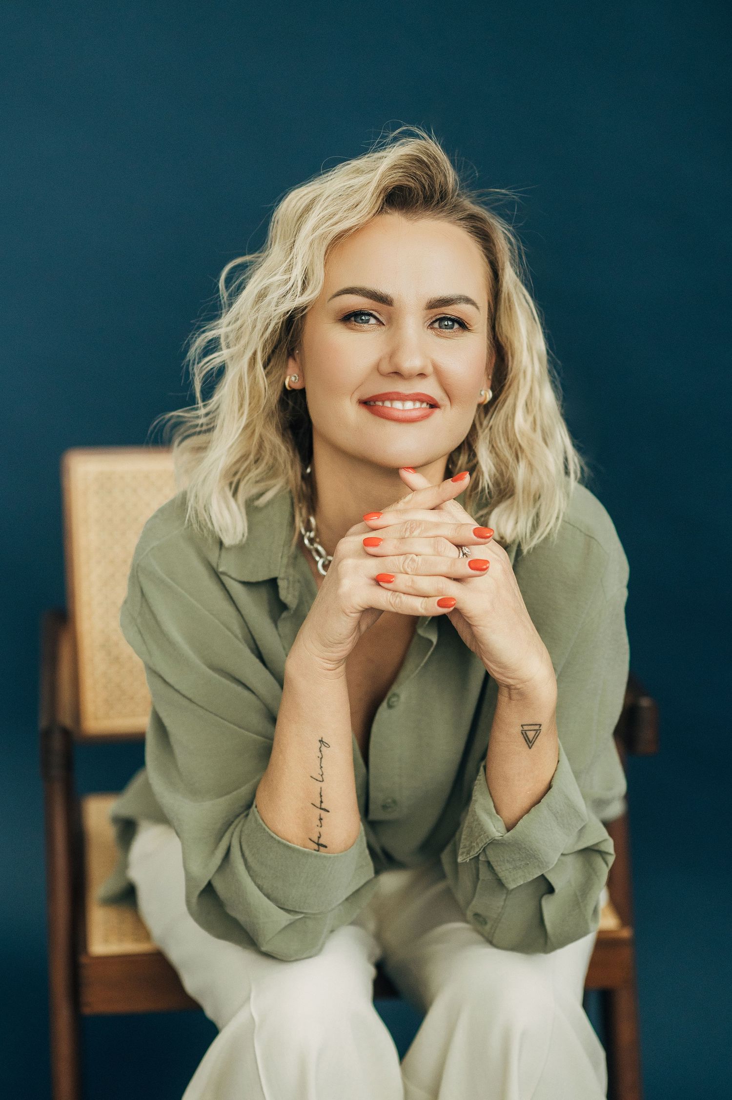 Professional studio portrait of someone in a sage green blouse sitting casually against a dark background.