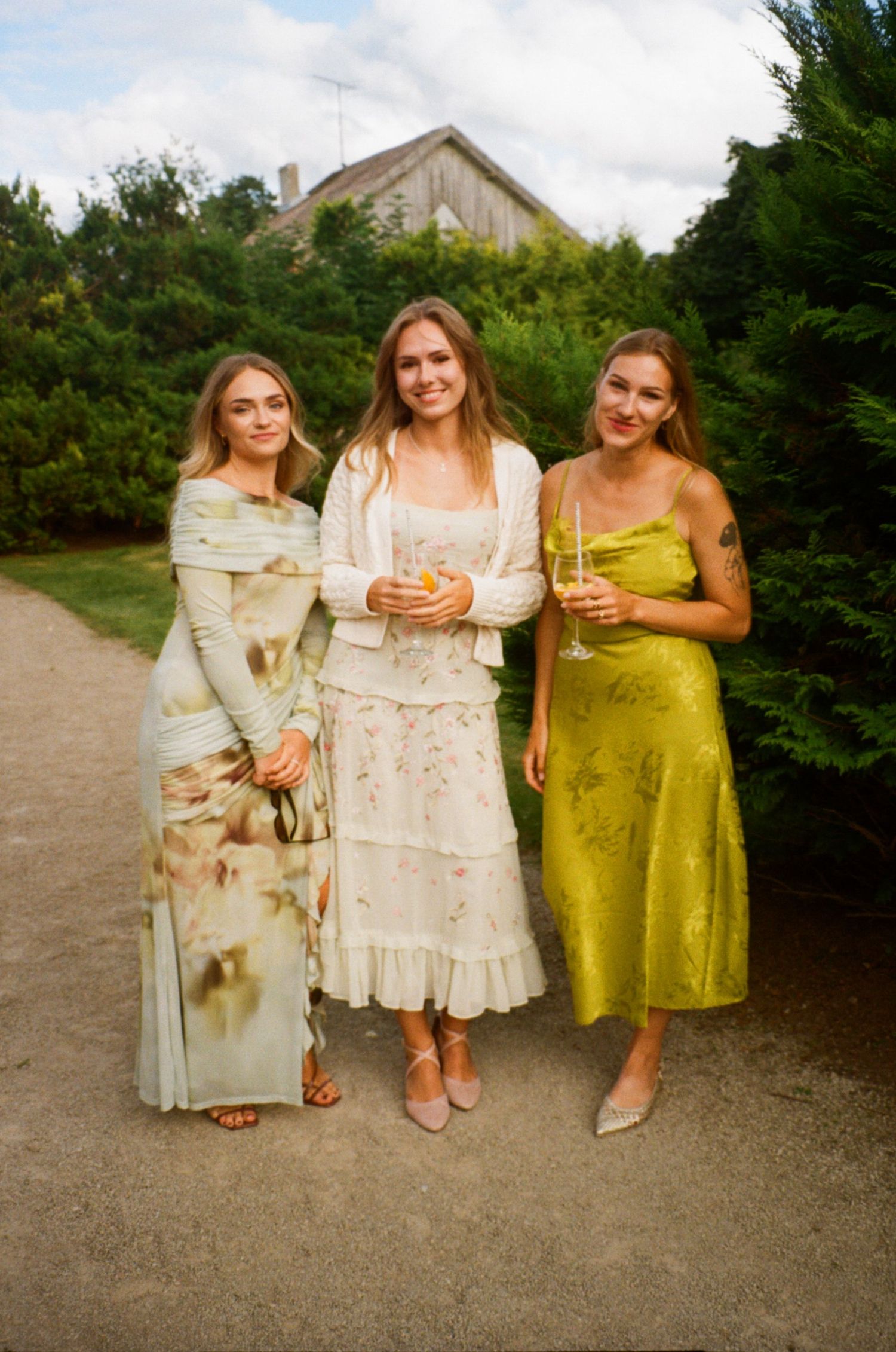 Three women in elegant dresses pose on a garden path, smiling and holding drinks at an outdoor event.