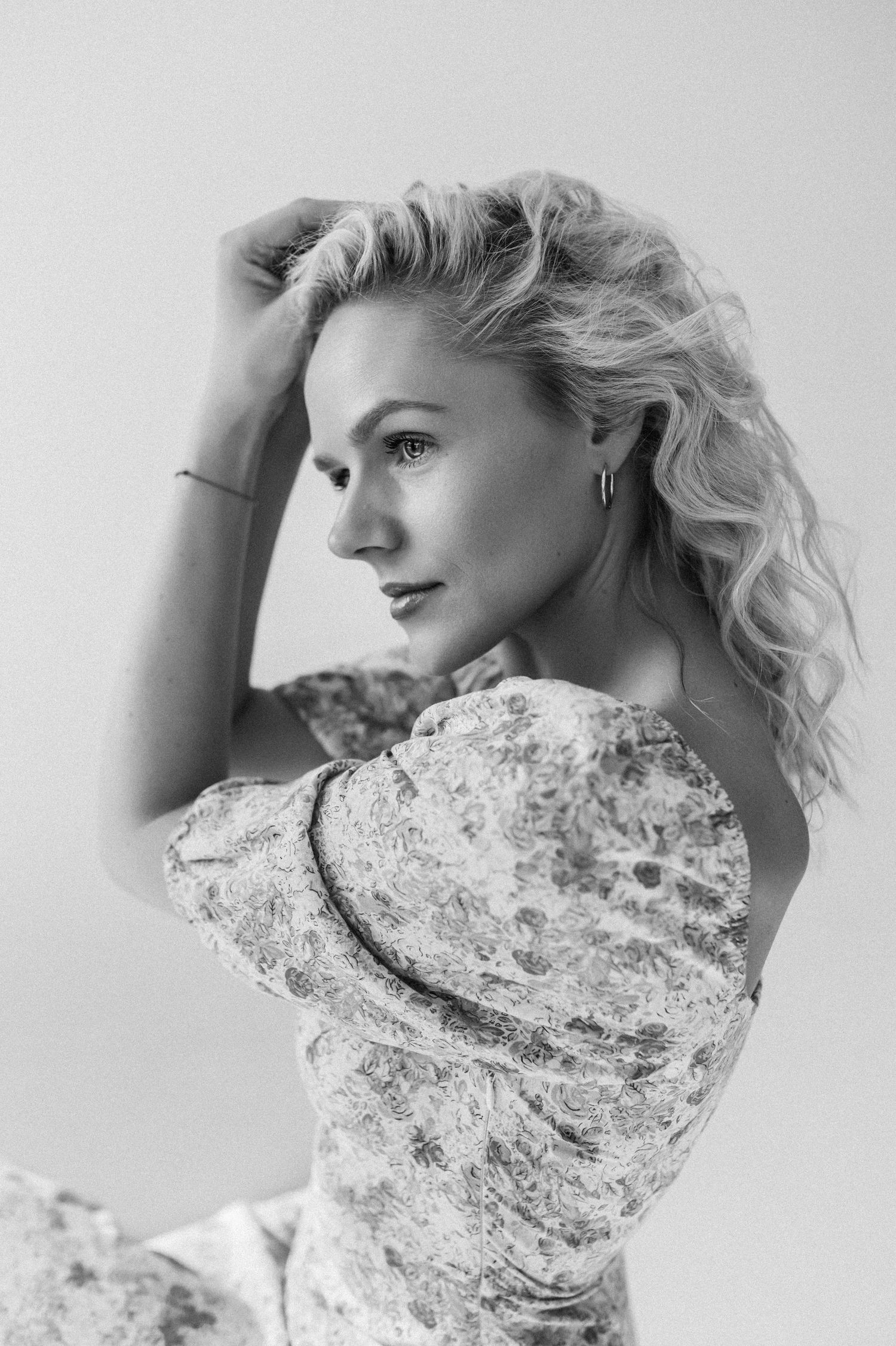 Black and white portrait of a blonde woman in a floral dress posing with hands in hair against a light background.