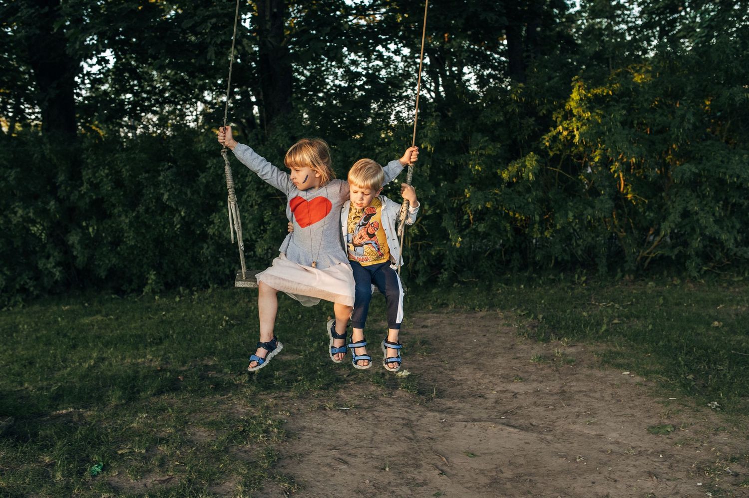 Two children sharing a wooden swing in a lush green park at dusk, smiling and playing together.