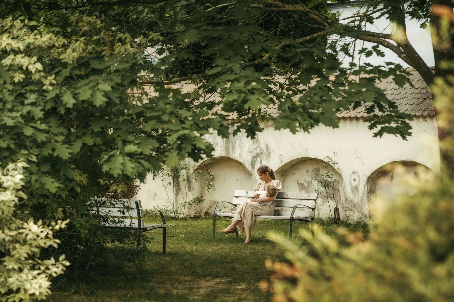 Woman sitting alone on a garden bench surrounded by lush green trees near a white arched wall.