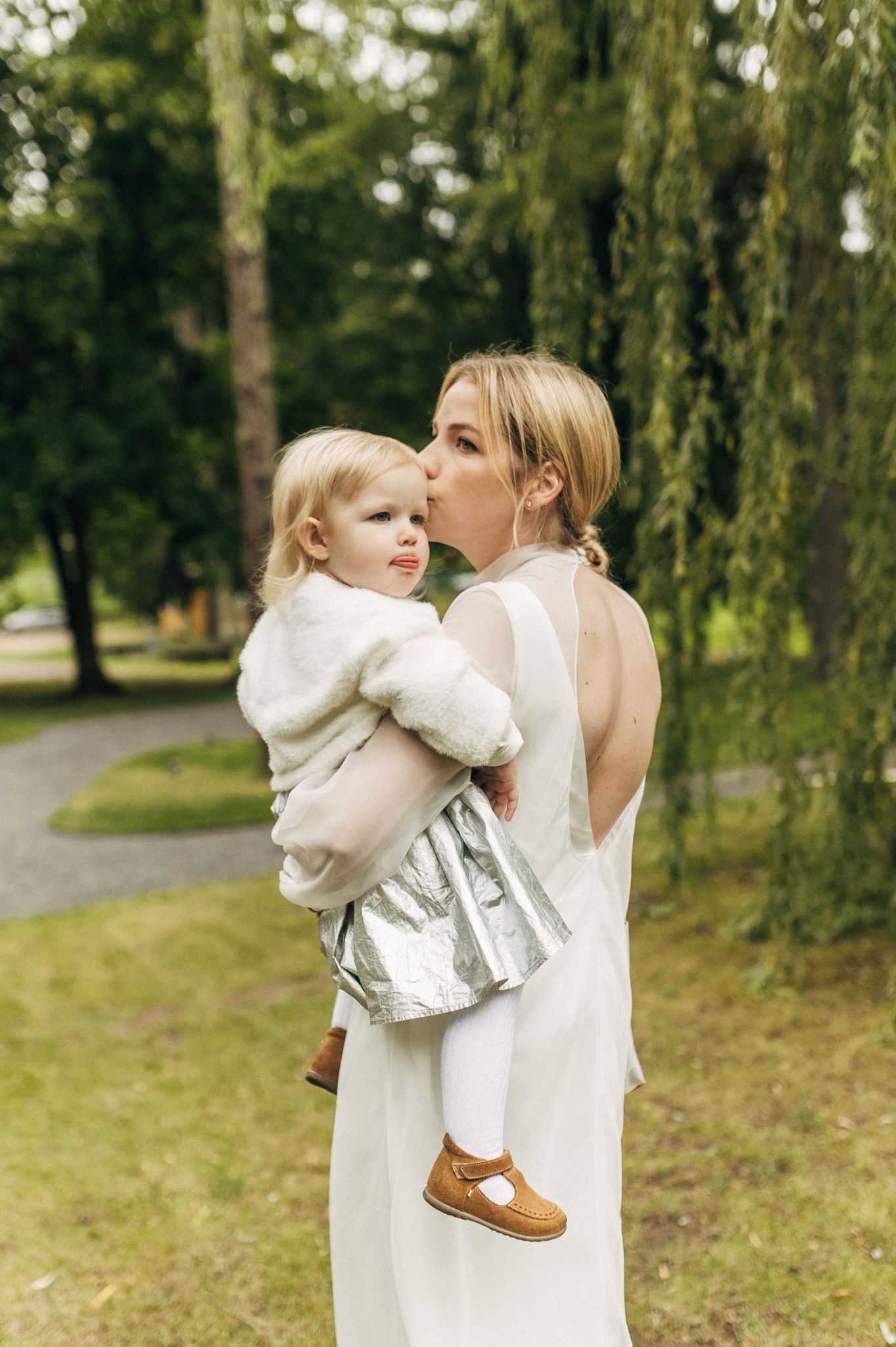 Bride in backless white gown kissing toddler daughter in park setting.