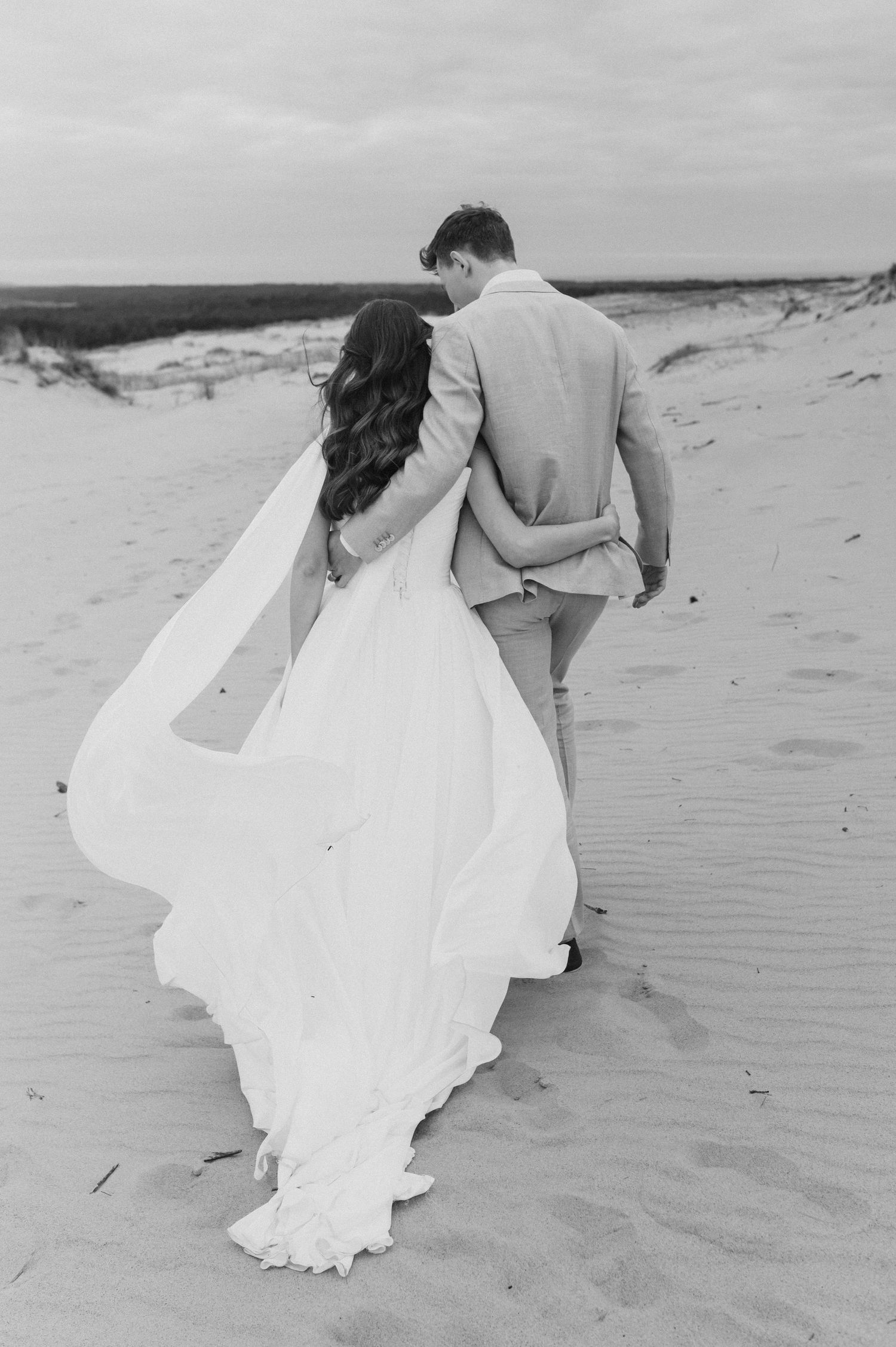 A flowing white dress billows in the wind as someone walks along a sandy beach in black and white photography.
