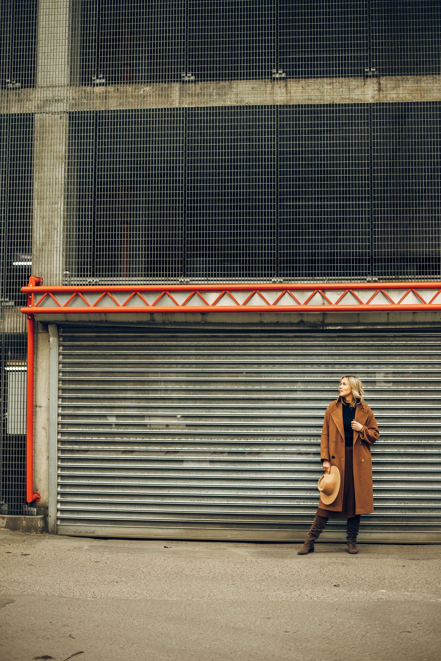Figure in brown coat posing against industrial metal shutters and concrete wall.