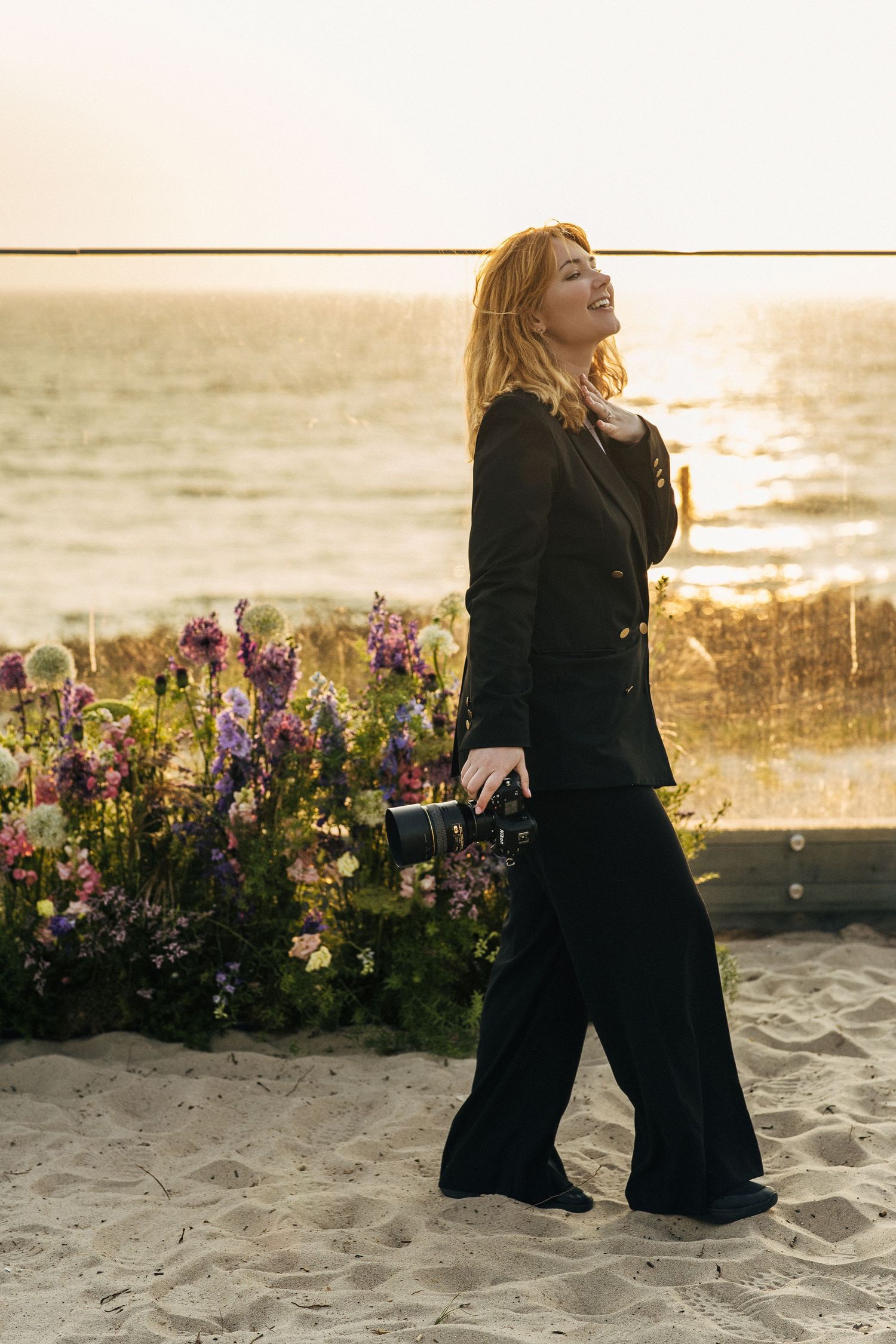 A person in a black outfit poses expressively on a sandy beach at sunset with purple wildflowers in the background.
