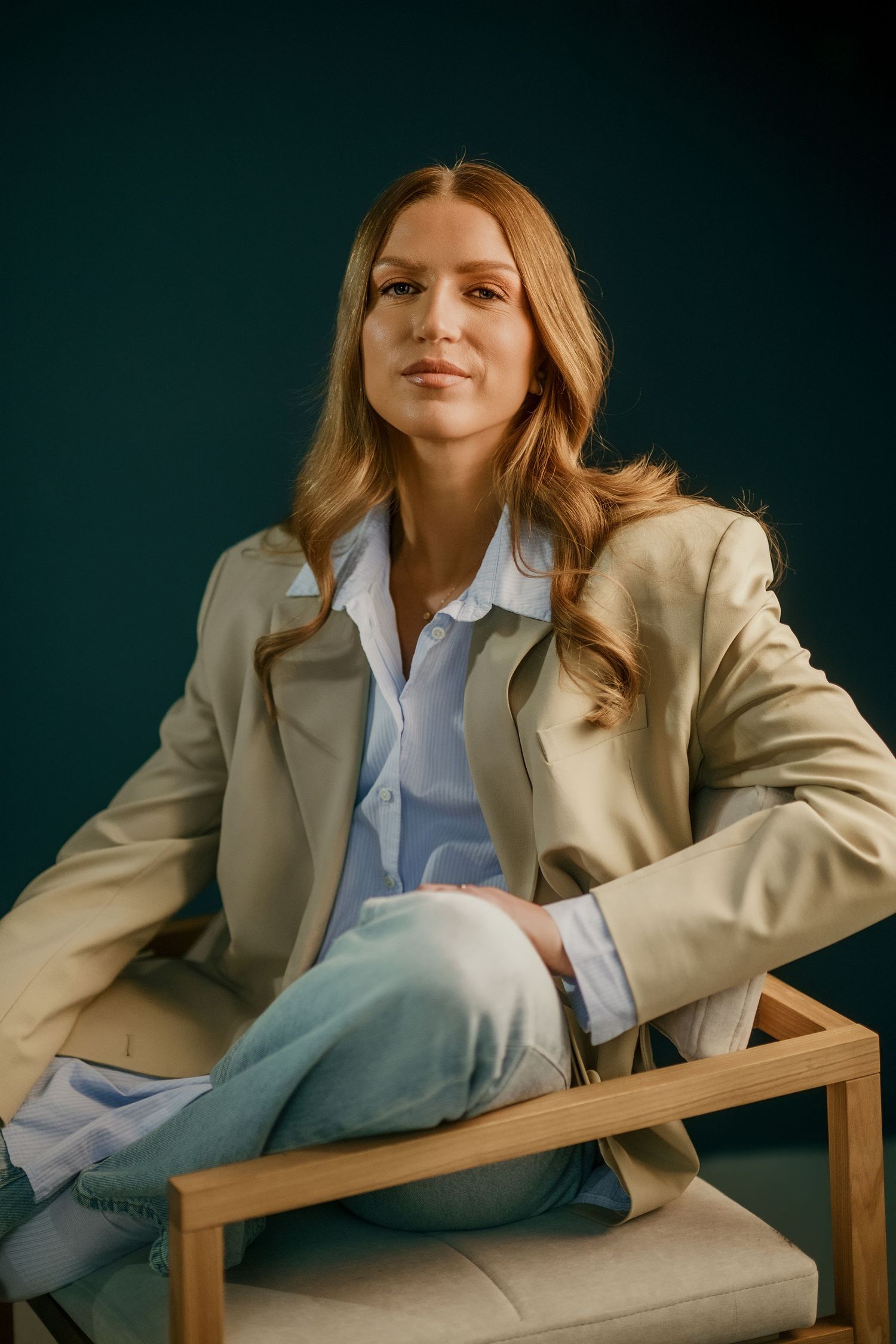 Professional wearing beige blazer and light blue shirt poses on modern chair against dark background.