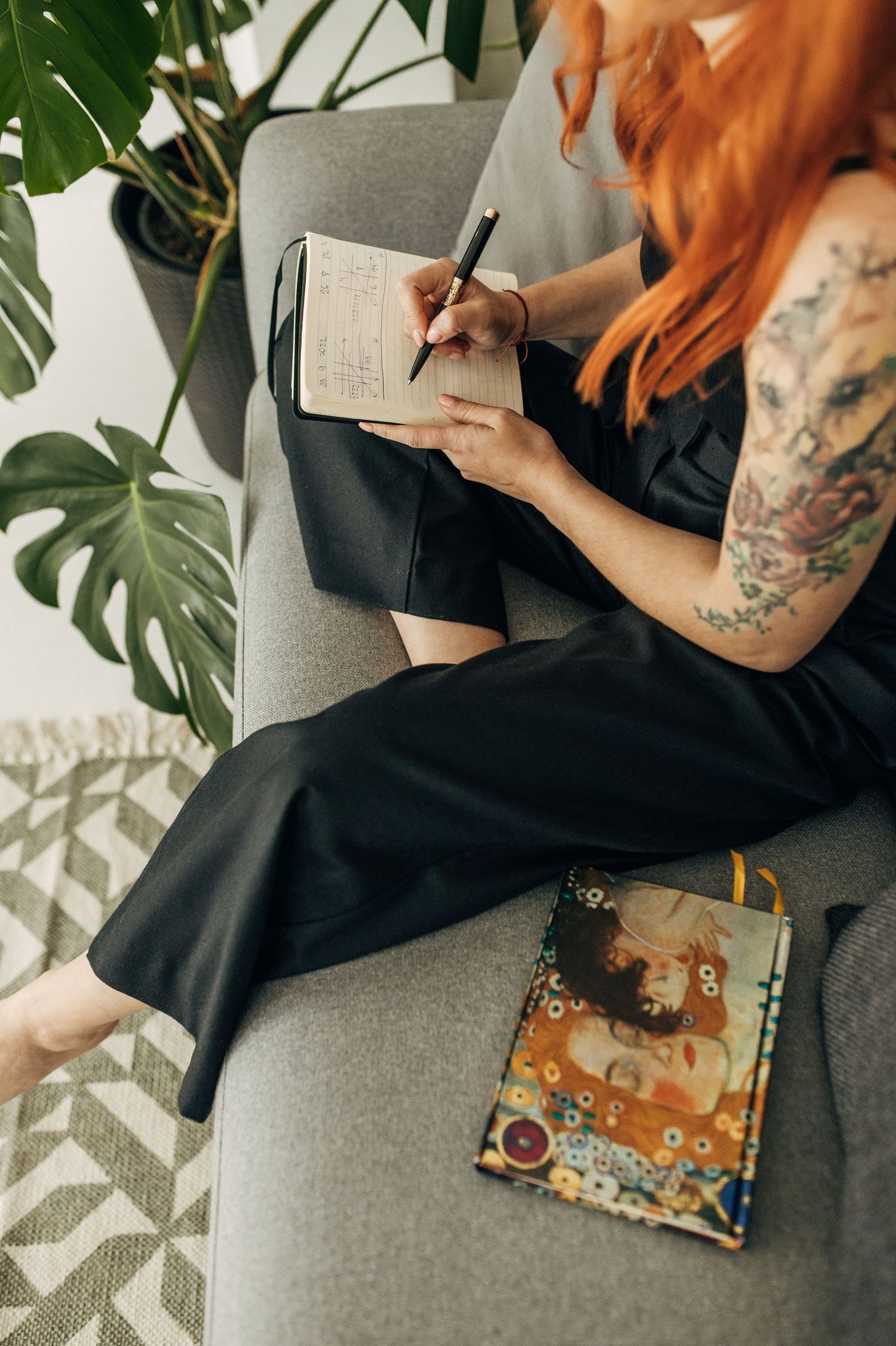 Tattooed hands writing in journal with tropical plants in background.