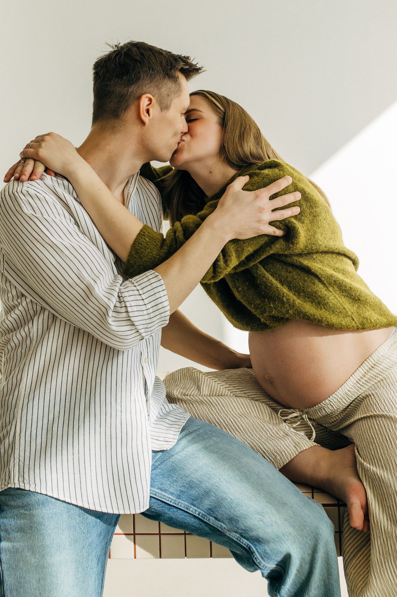 Pregnant woman in green sweater kissing her partner while sitting, showing baby bump in a bright studio setting.
