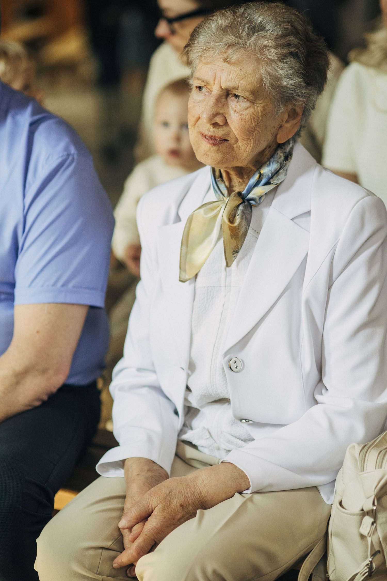 Elegant elderly woman in white blazer with silk scarf seated at a formal gathering.