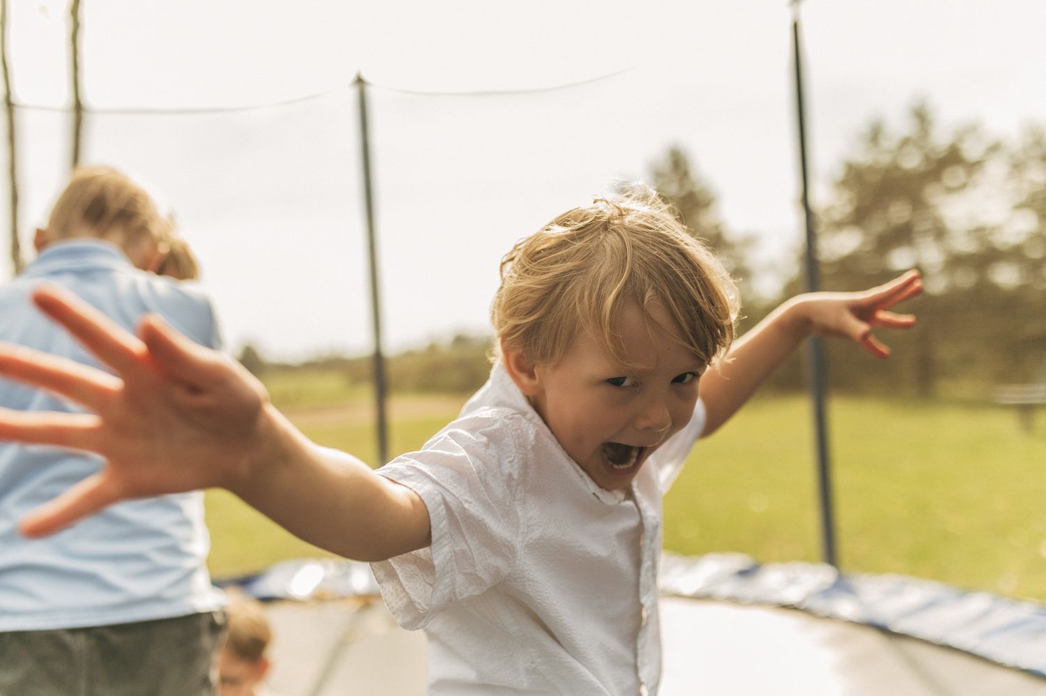 Excited blonde toddler boy jumping on a trampoline outdoors with arms outstretched.