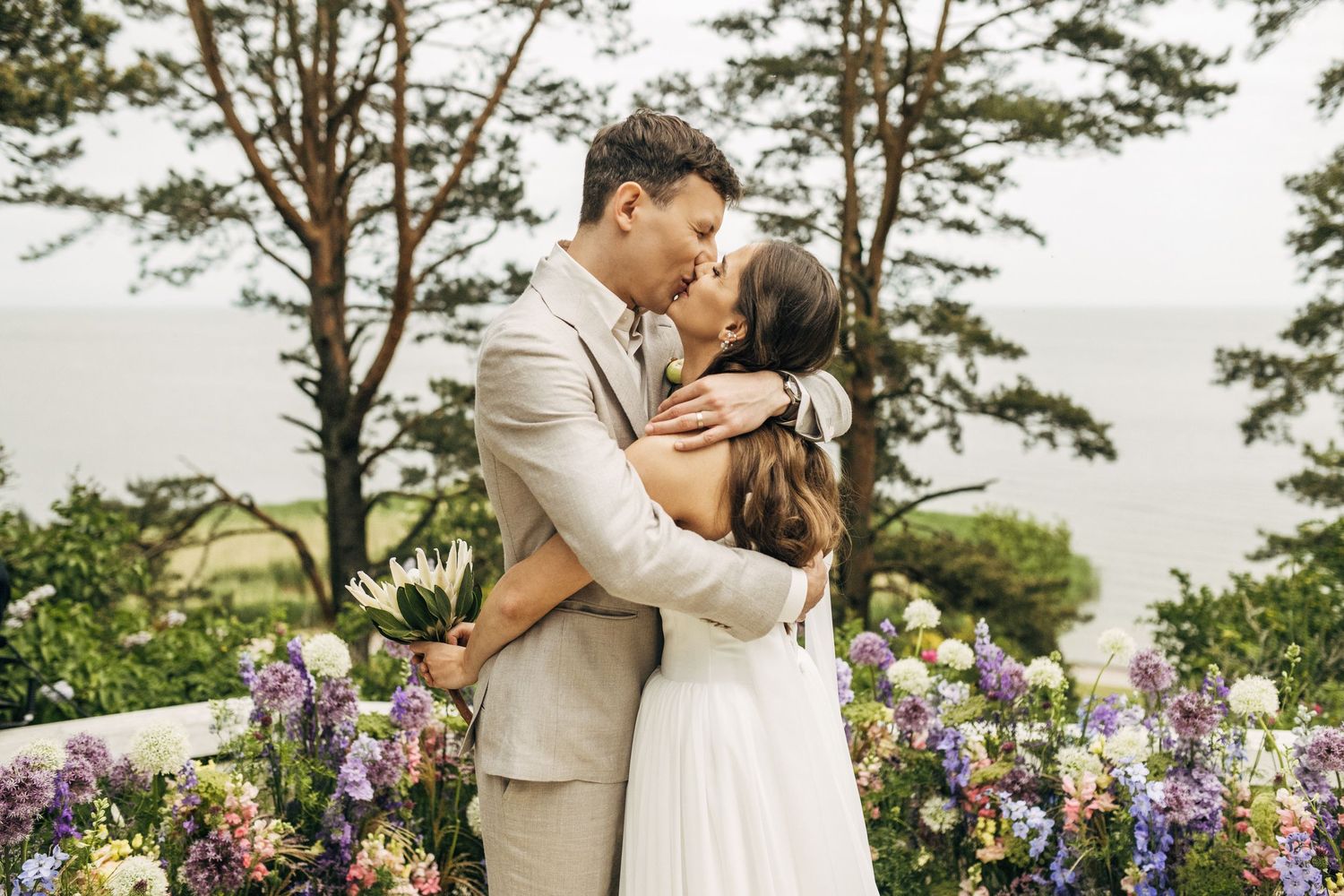 A romantic wedding couple shares a kiss in a garden overlooking the ocean with purple flowers in the foreground.