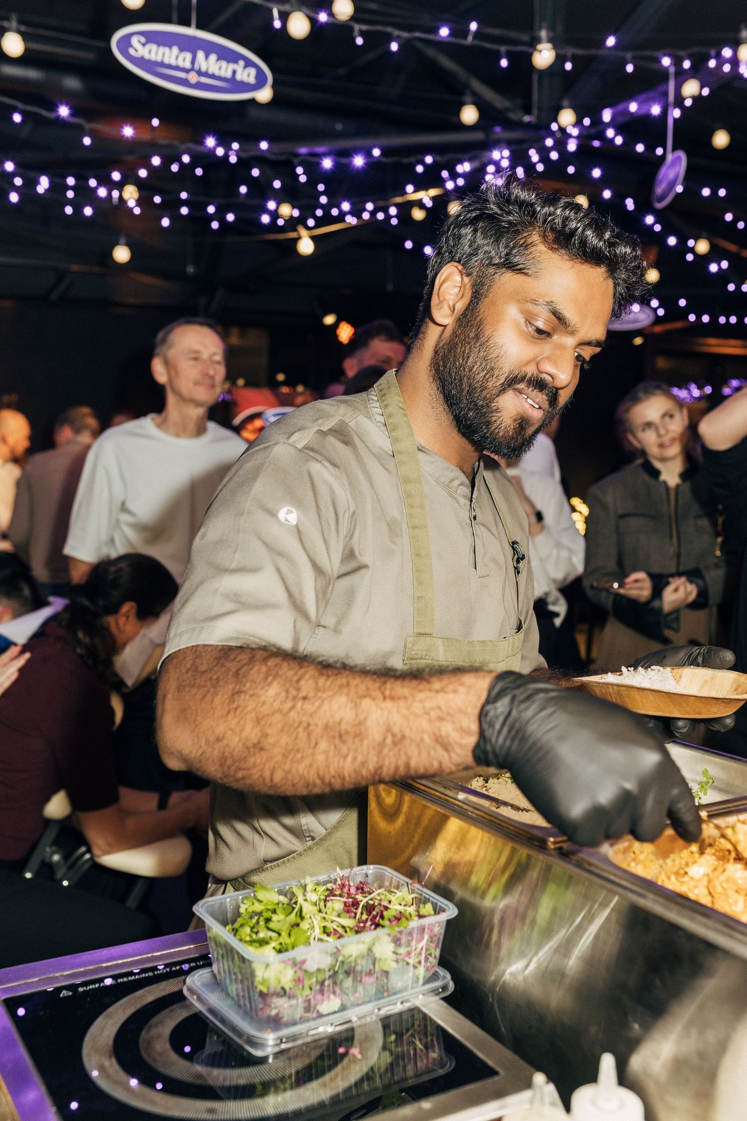 Chef preparing fresh food at an outdoor event under twinkling string lights and purple ambient lighting.