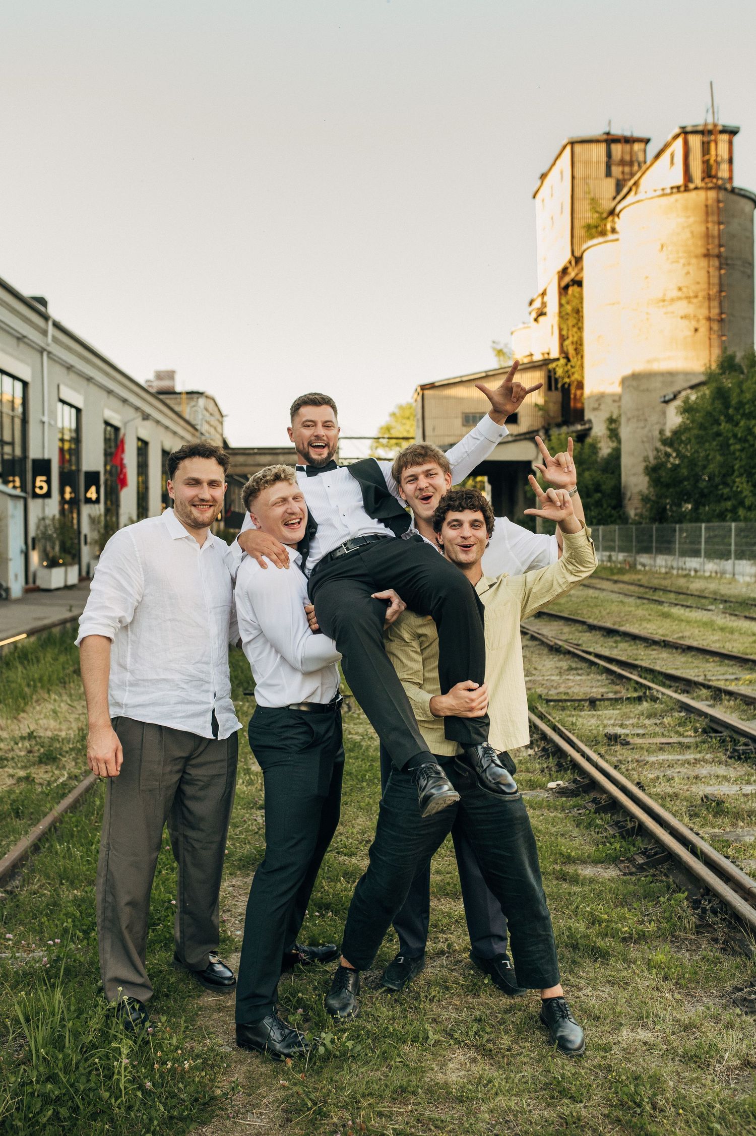 Group of five young men celebrating outdoors on railway tracks, one being lifted up, near industrial buildings.