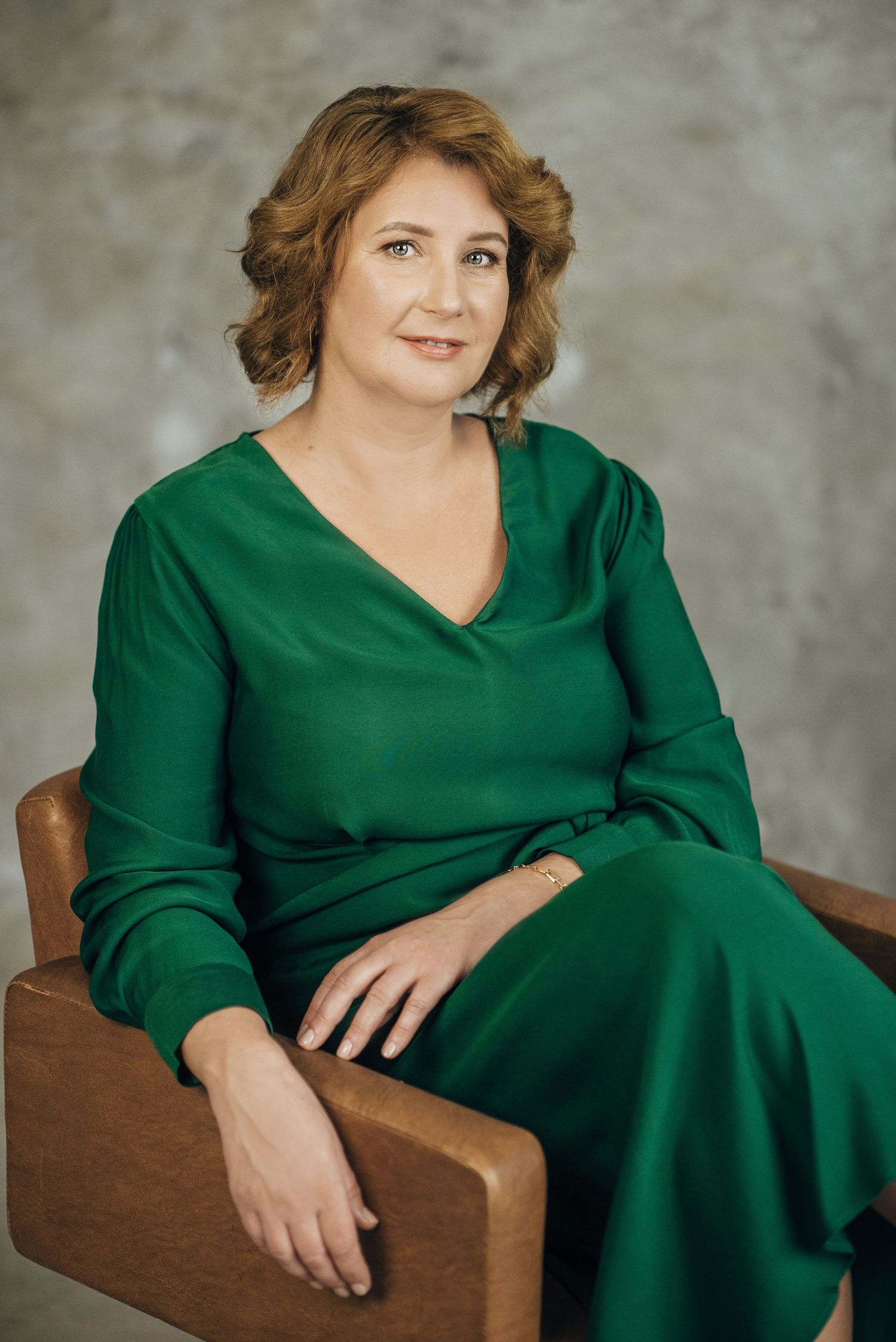 Portrait series in emerald green dress seated in brown leather chair against textured wall.