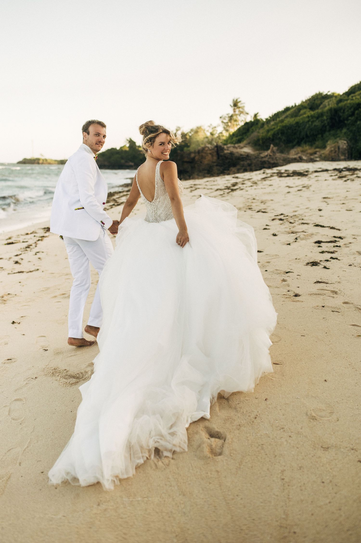 A couple in wedding attire walks along a sandy beach at sunset creating romantic silhouettes.