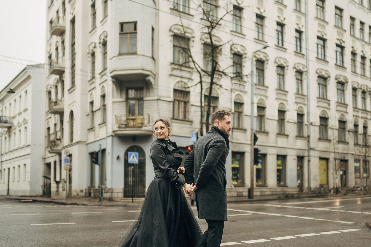 A couple poses playfully on a city street corner with historic architecture in the background.