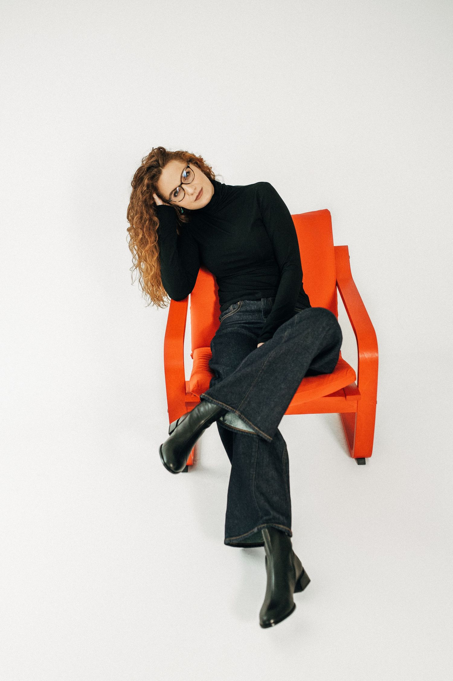 Stylish portrait of person in black turtleneck and jeans lounging on orange chair.