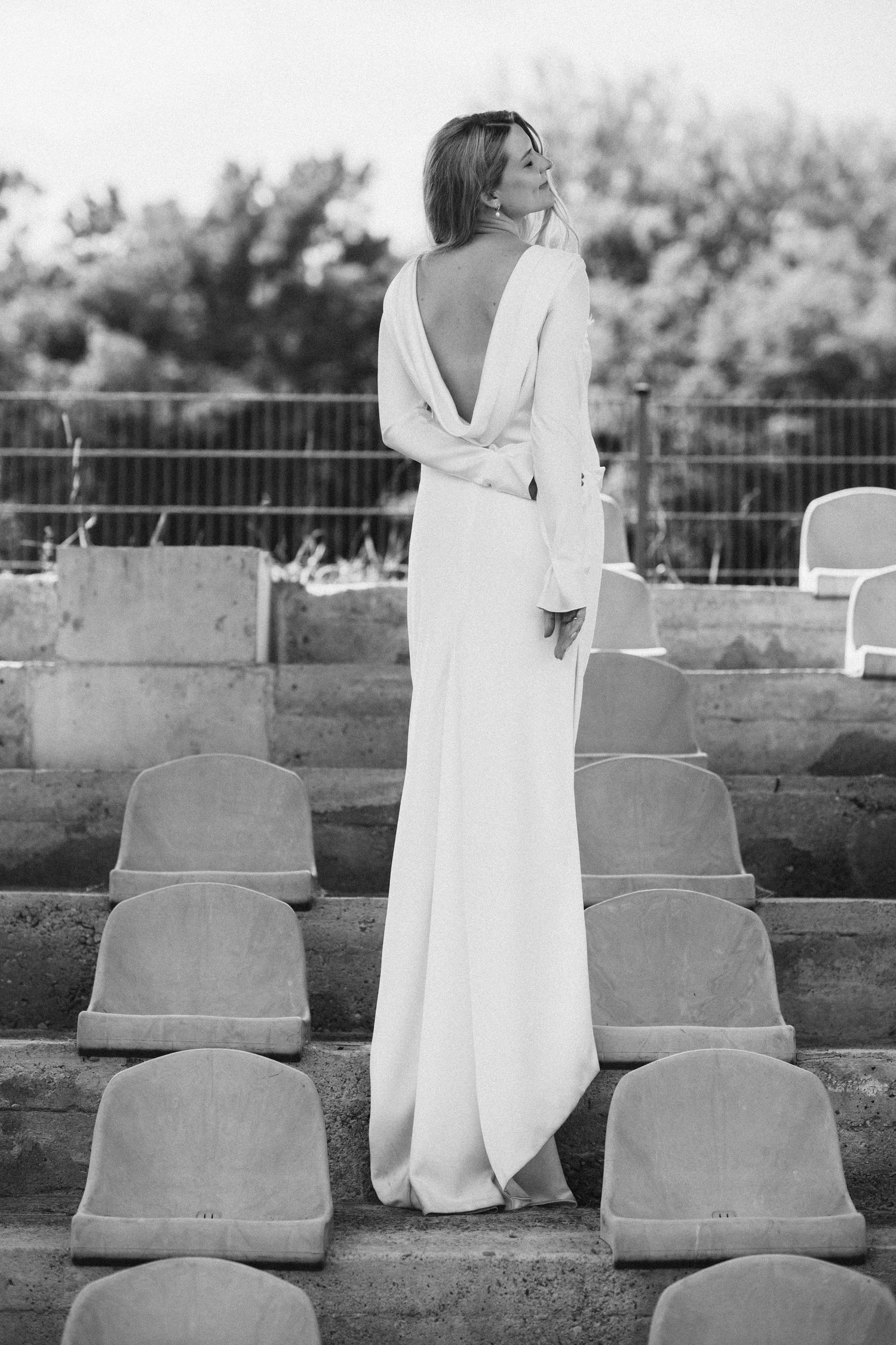 Black and white photo of a woman in an elegant backless white gown standing among stadium seats outdoors.