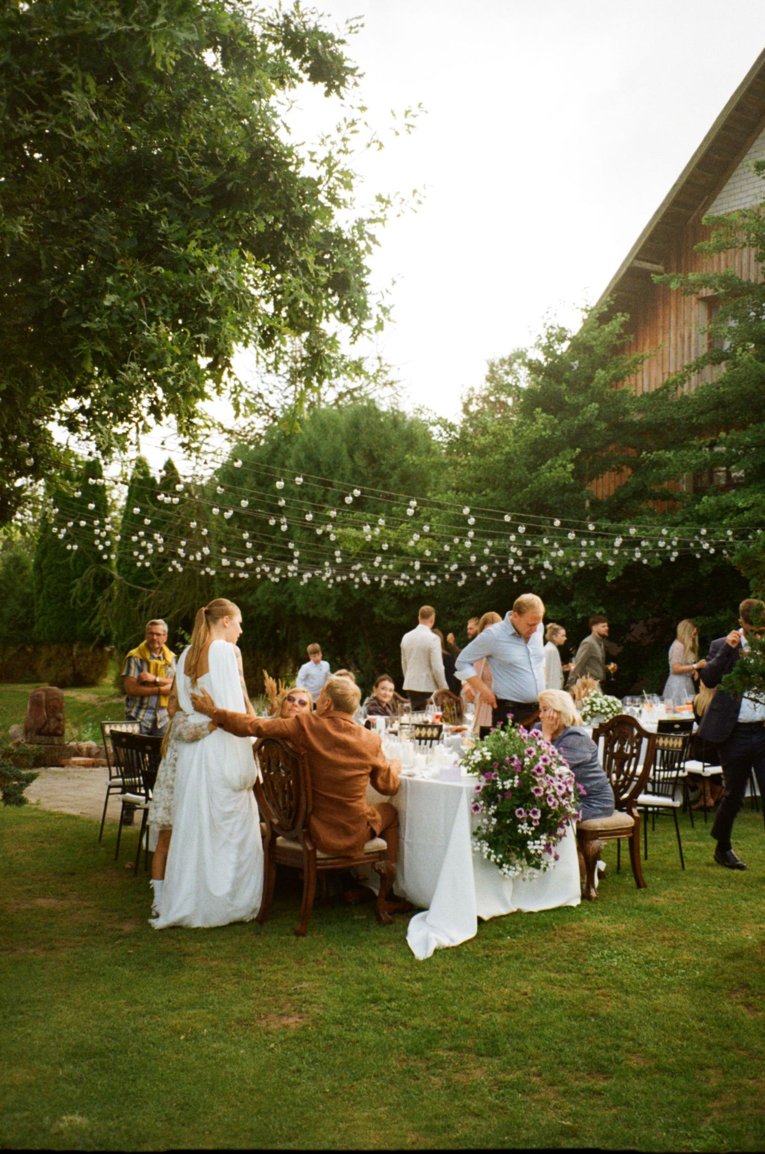 Outdoor wedding reception on green lawn with string lights, floral table, bride in white dress, and guests dining at sunset.