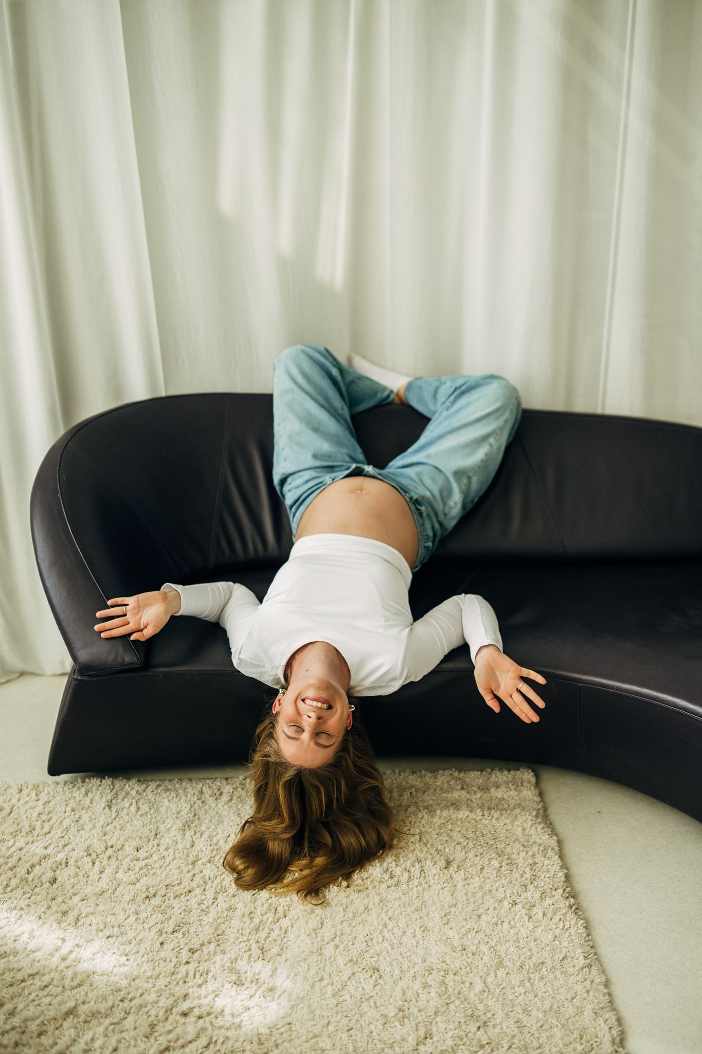 Young woman in jeans and white top lying upside down on a dark leather sofa, smiling and relaxed at home.