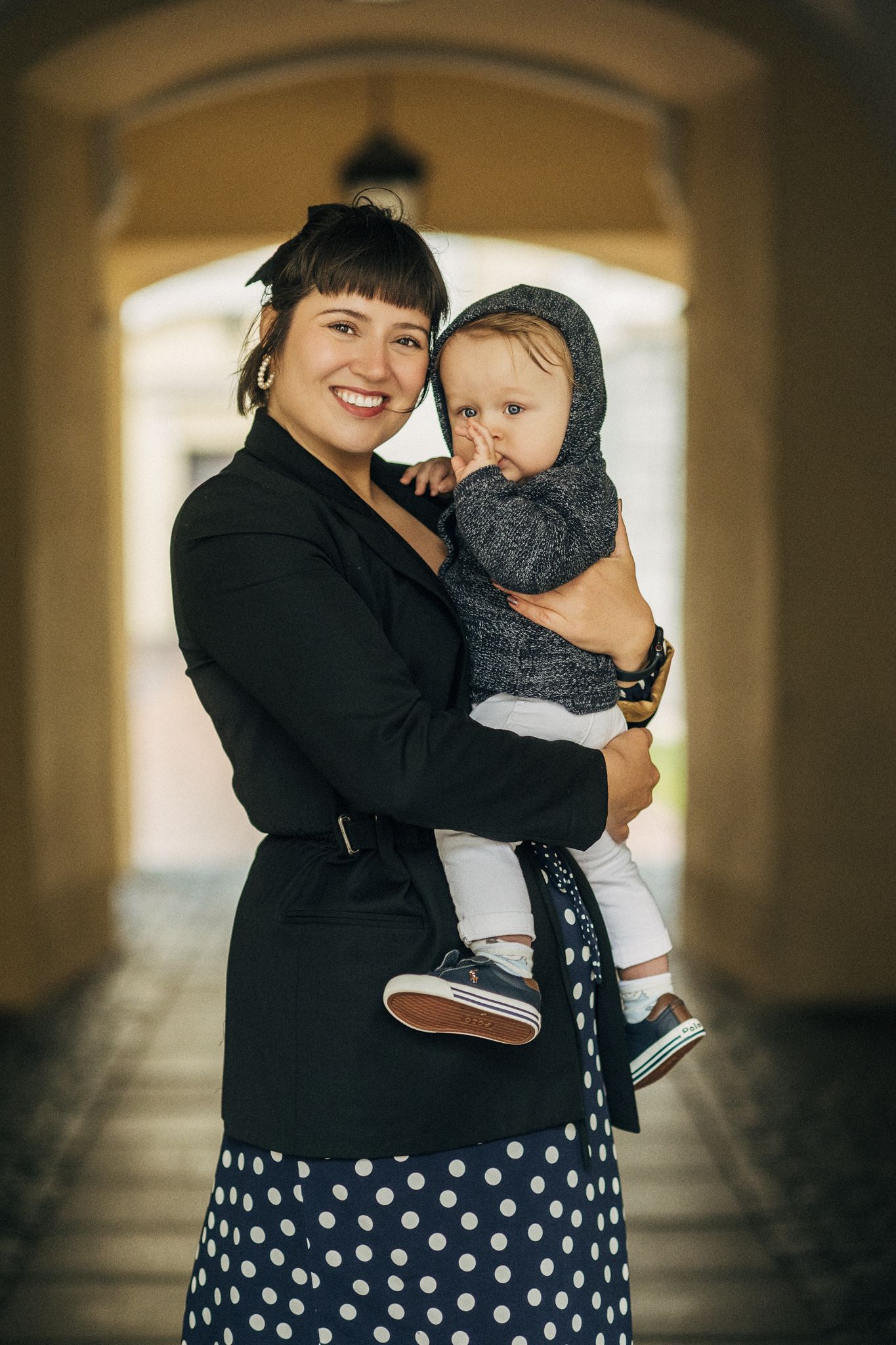 A smiling woman in a black blazer and polka dot skirt holds a baby in a gray hat.