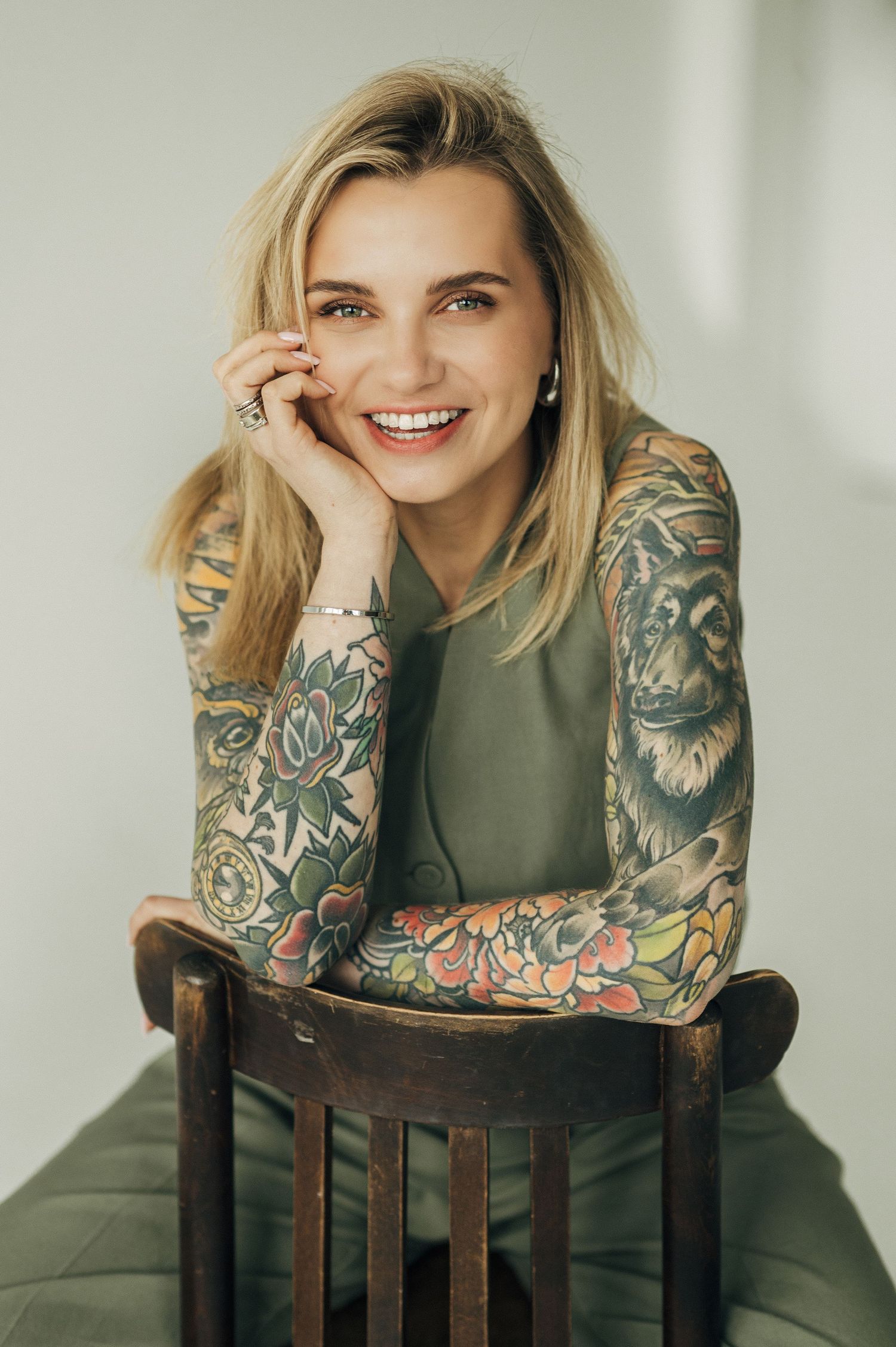 Model with colorful sleeve tattoos poses on wooden chair in olive green outfit against light background.