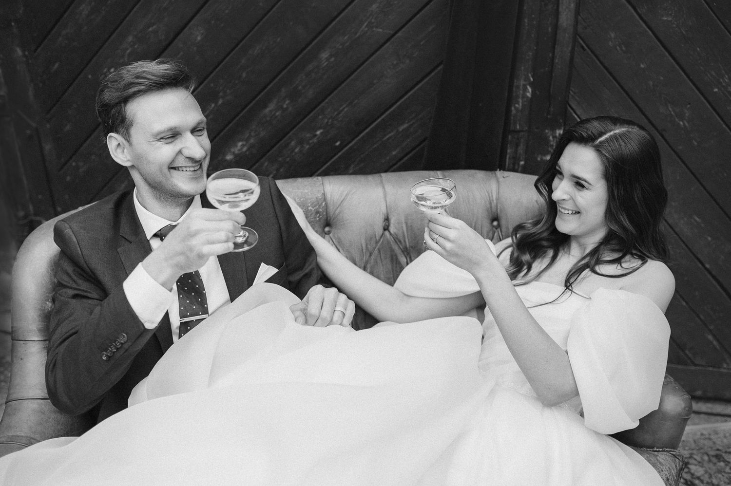 A couple in wedding attire laughing together while seated on an elegant tufted couch.