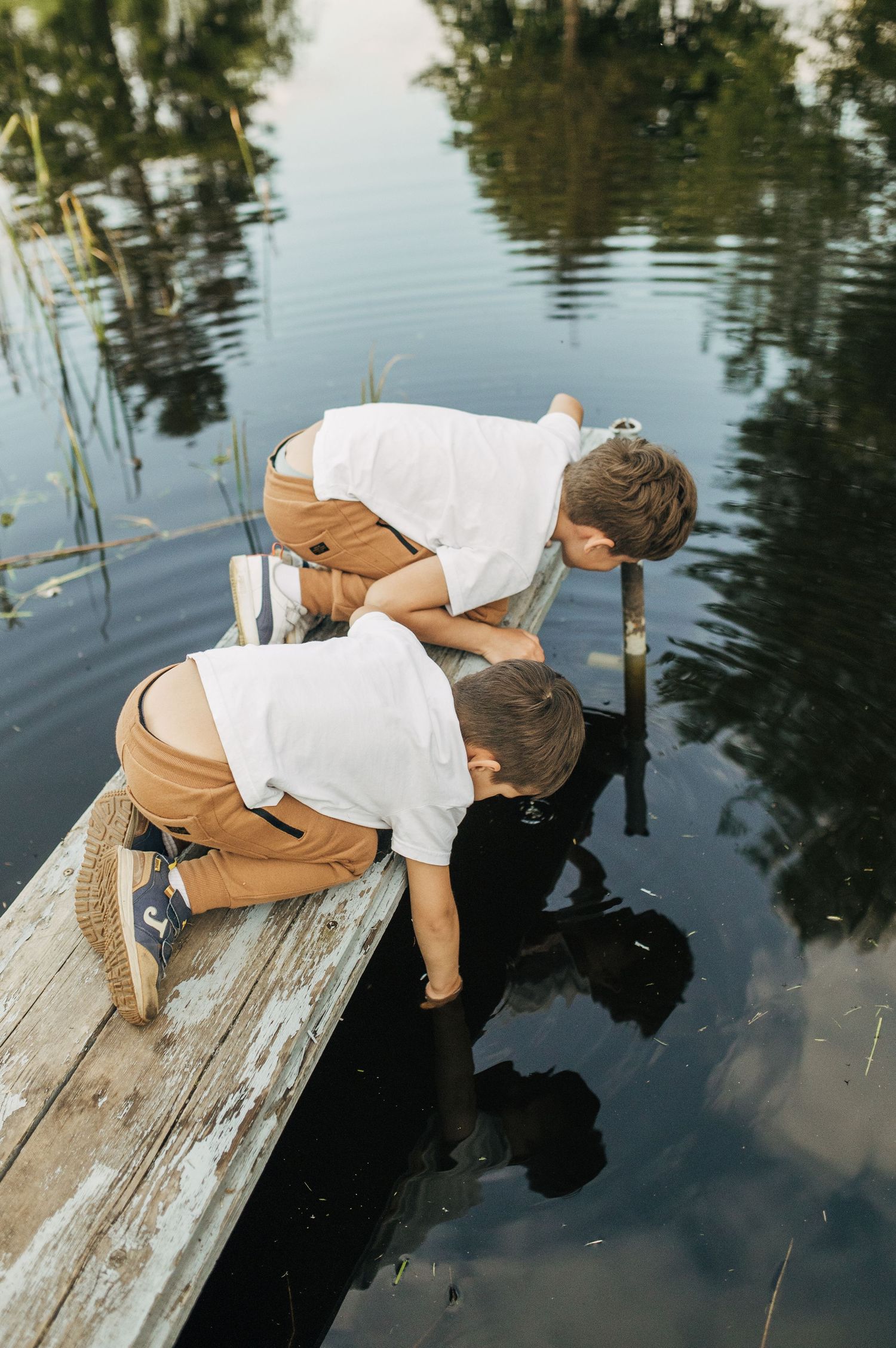 Child leans over wooden dock to look at reflection in still water wearing white and tan casual clothes.