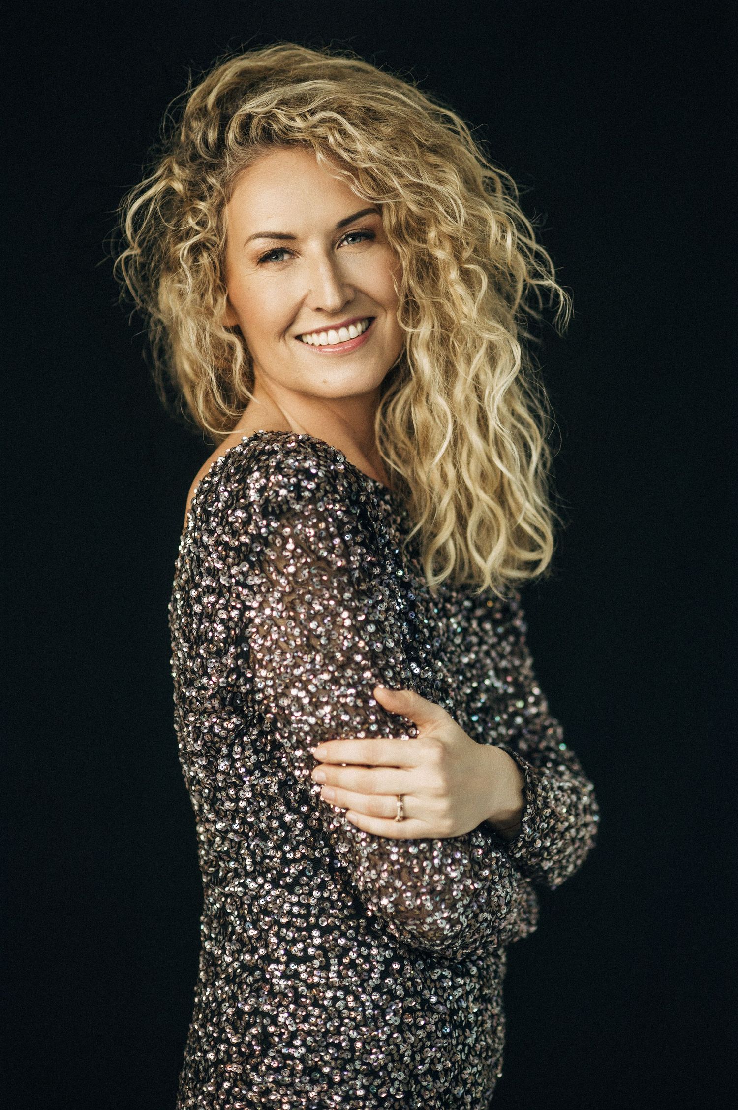 Smiling woman with curly blonde hair wearing a sparkly sequin dress poses against a dark background.