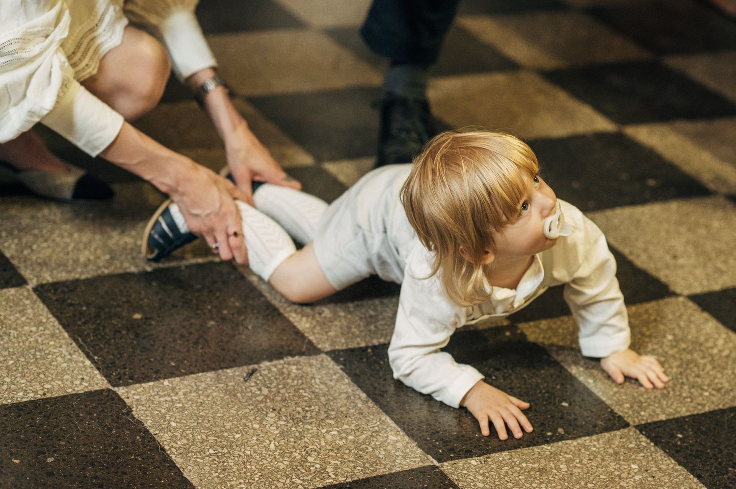 Blonde toddler crawling on a checkered floor during a formal event with adults nearby.