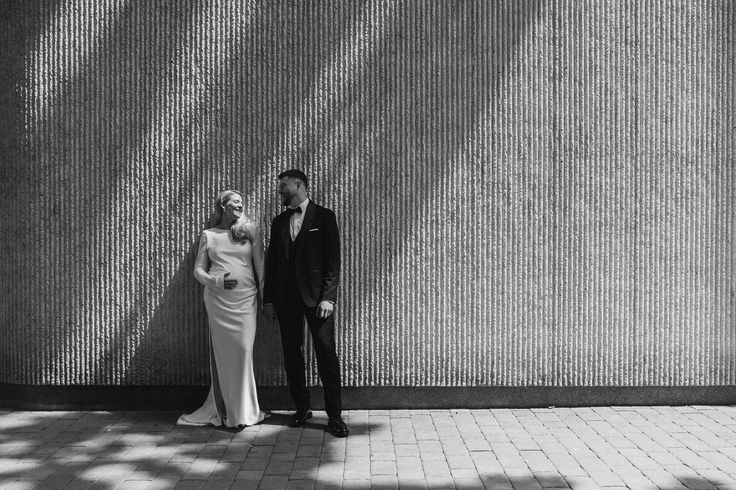 Black and white photo of a bride and groom posing against a textured wall with dramatic shadow patterns.