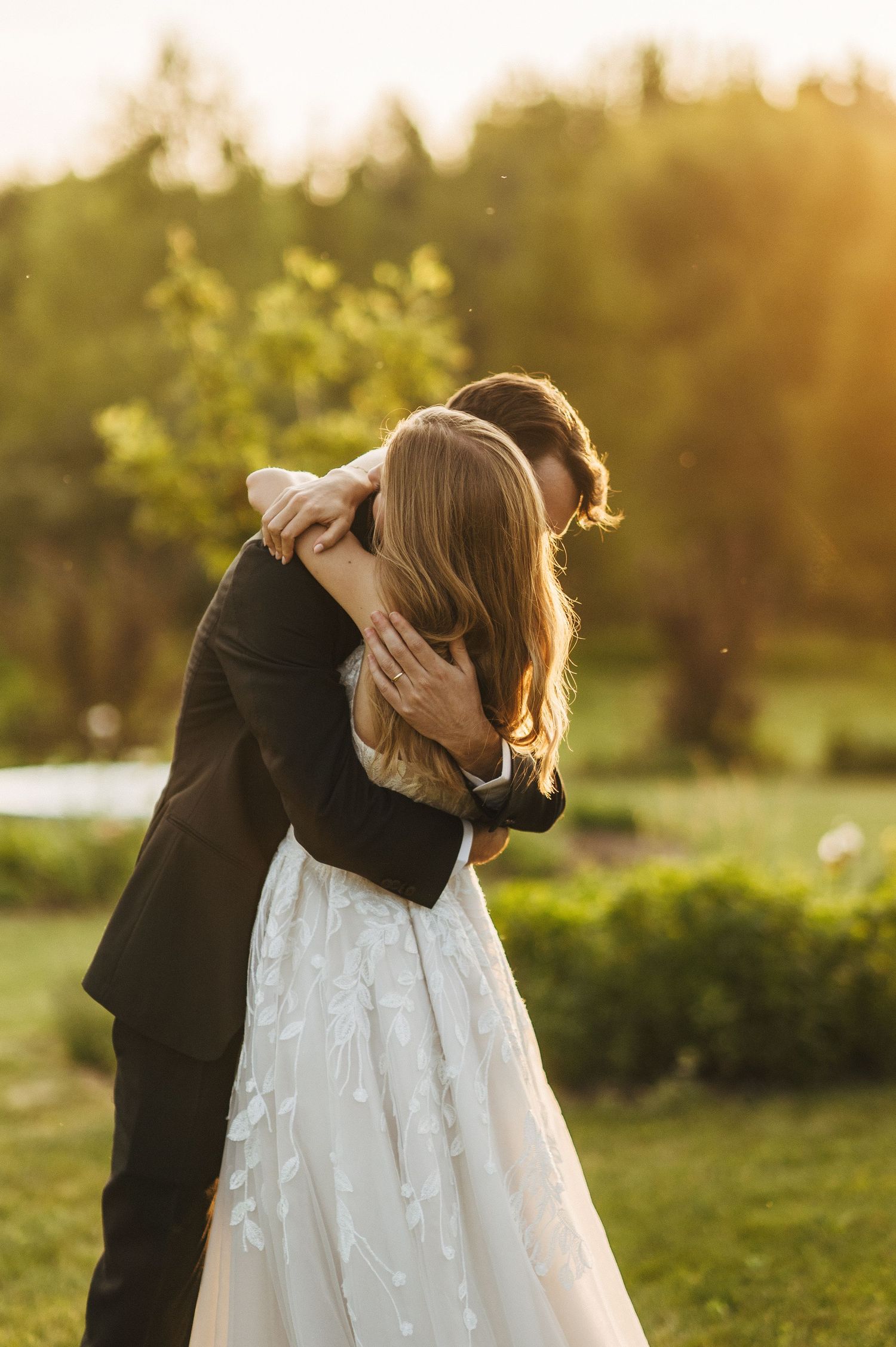A bride and groom share a romantic embrace in golden sunset light, surrounded by lush green gardens on their wedding day.