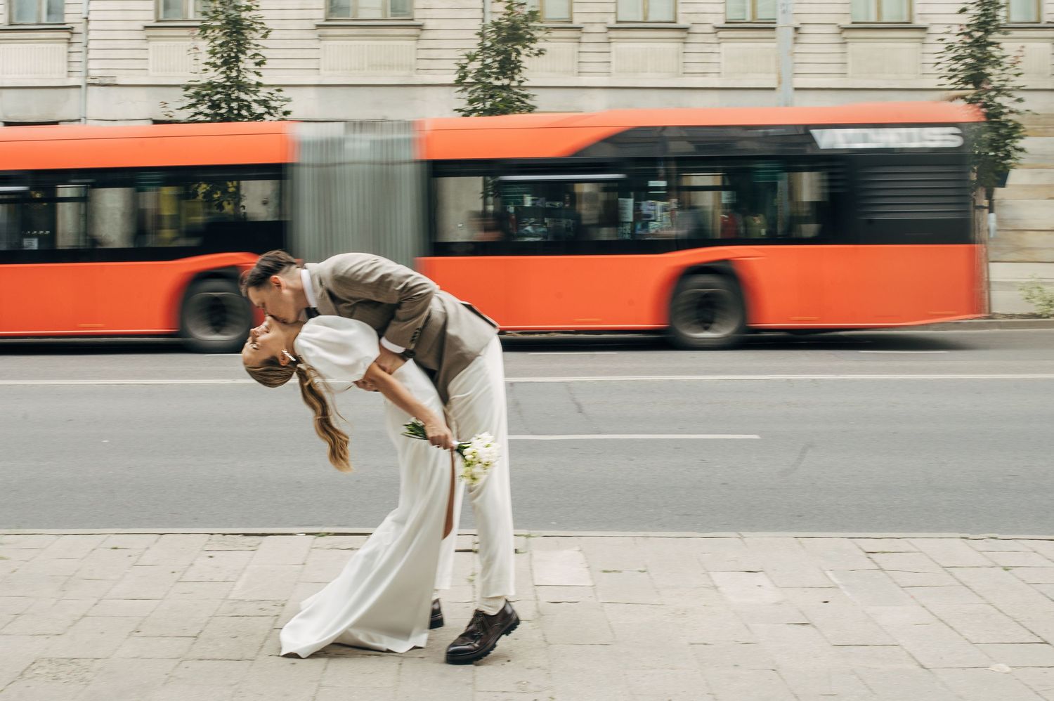 Newlyweds embrace in a dip pose on a city street with an orange bus passing by.