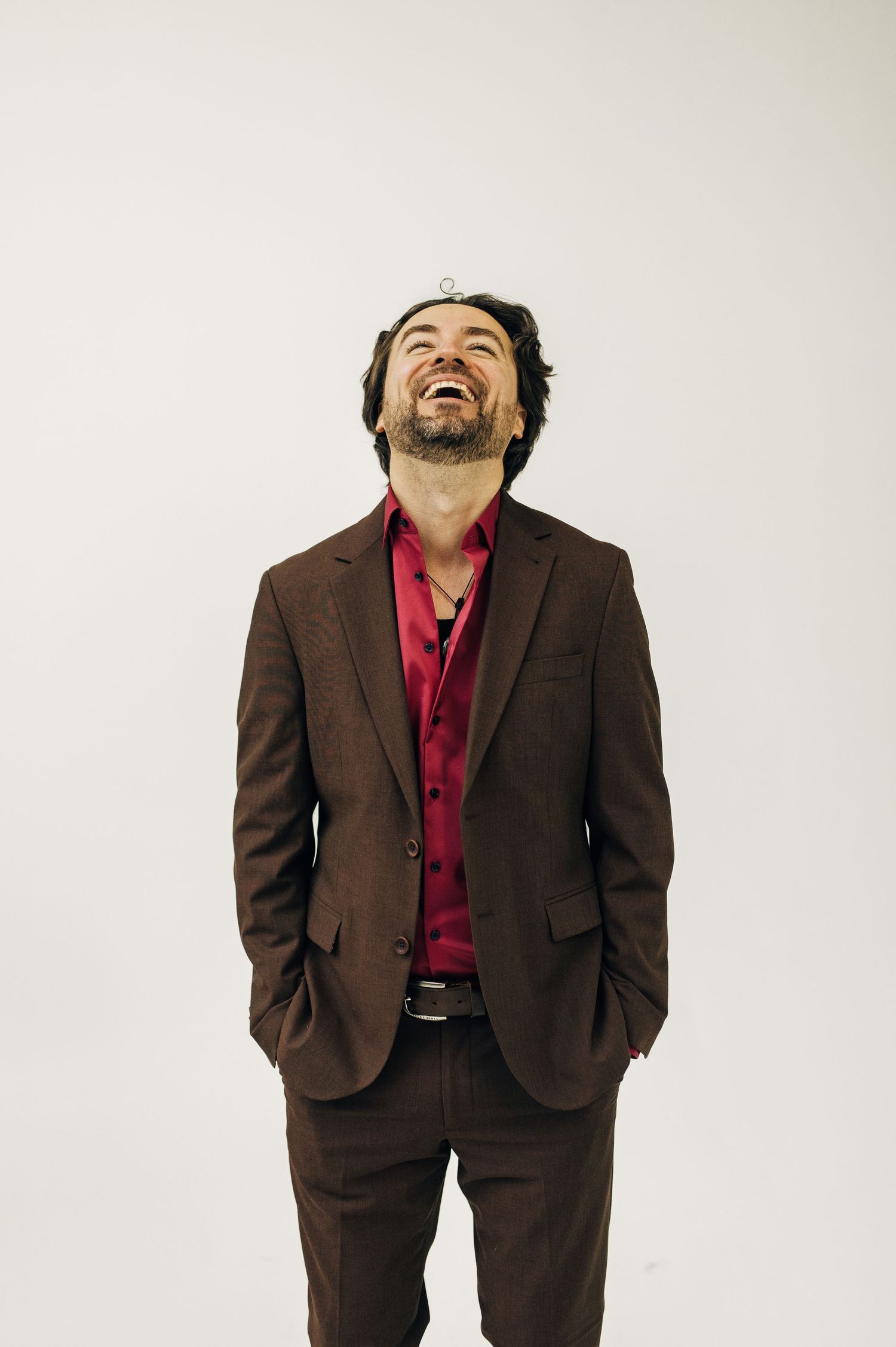 A laughing man in a brown suit and red shirt stands confidently with hands in pockets against a white background.