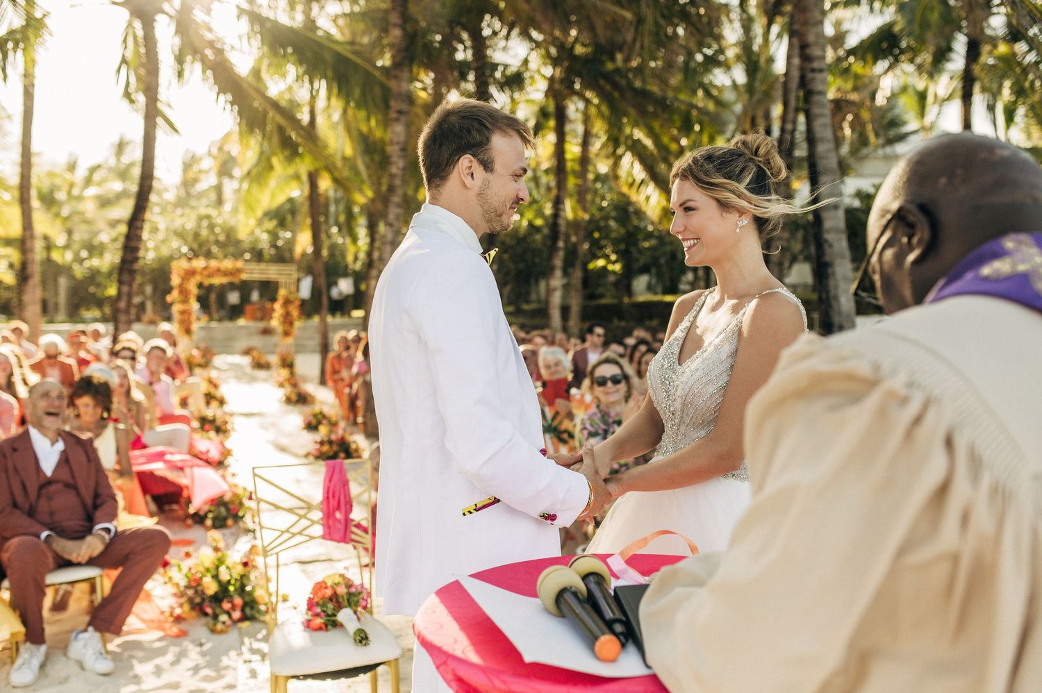 A couple holds hands during their outdoor wedding ceremony surrounded by palm trees and red decor.