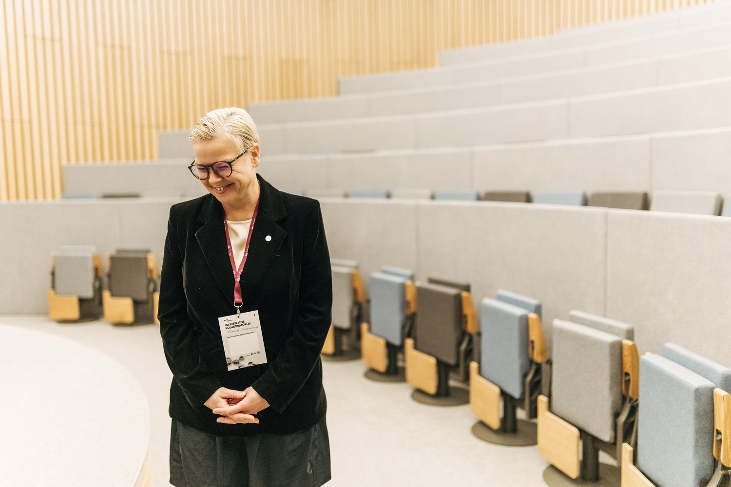 A person in a black blazer stands in a modern lecture hall with tiered seating and wooden desks.