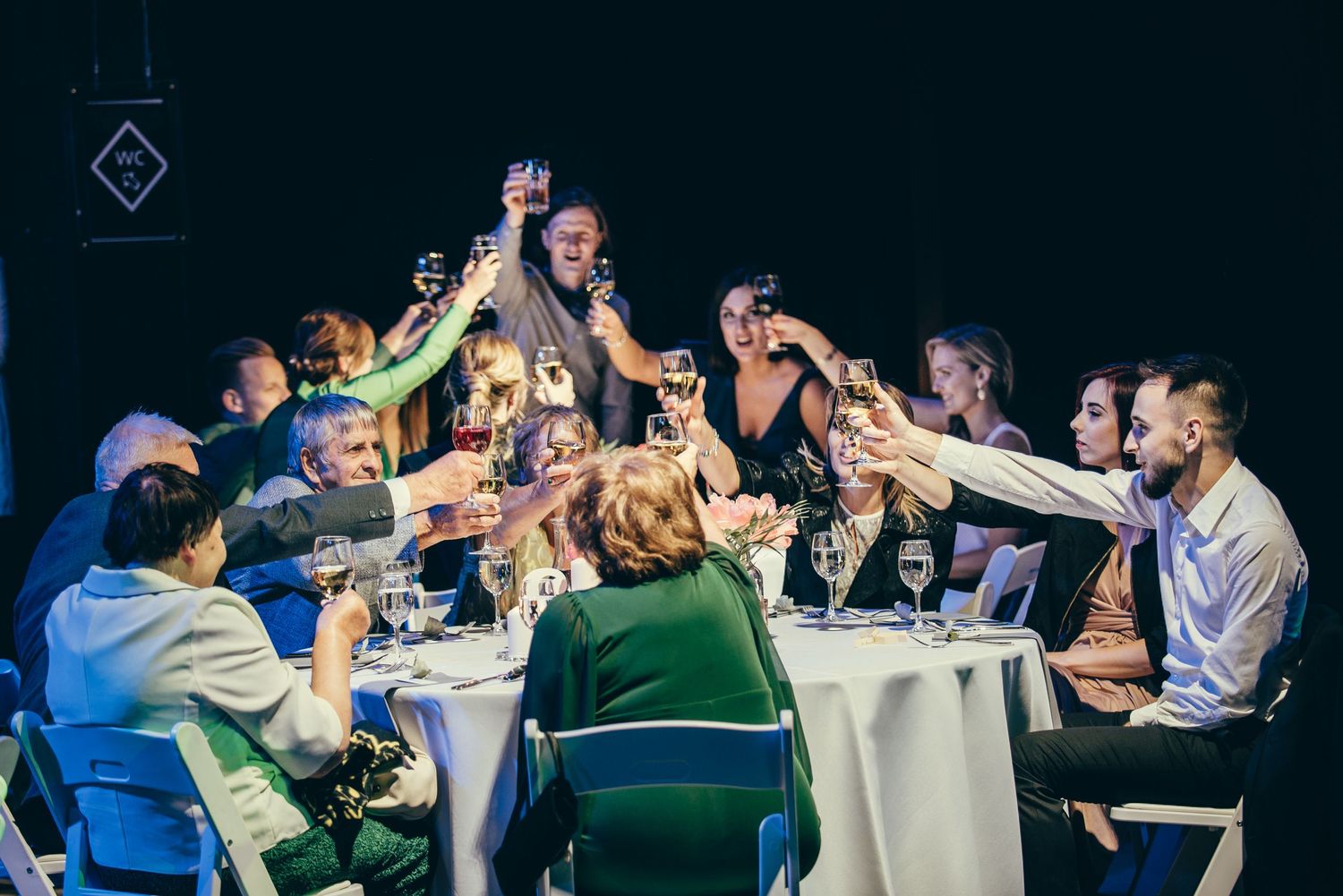 Wedding reception guests raising glasses in celebration at tables lit by ambient lighting.