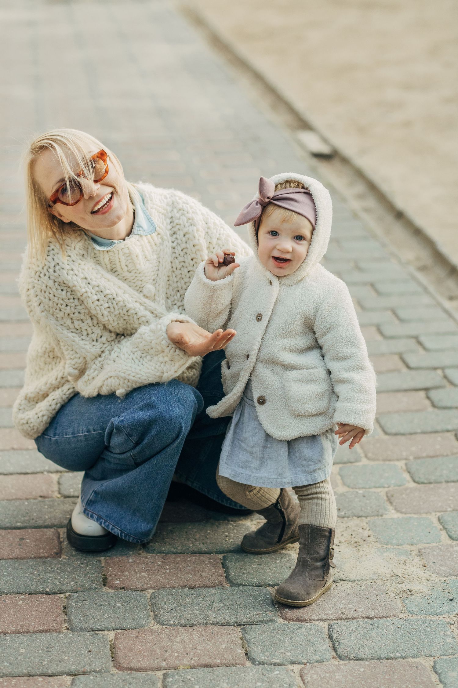 Candid sequence showing parent and toddler interacting on brick pavement while wearing cream-colored knitwear.