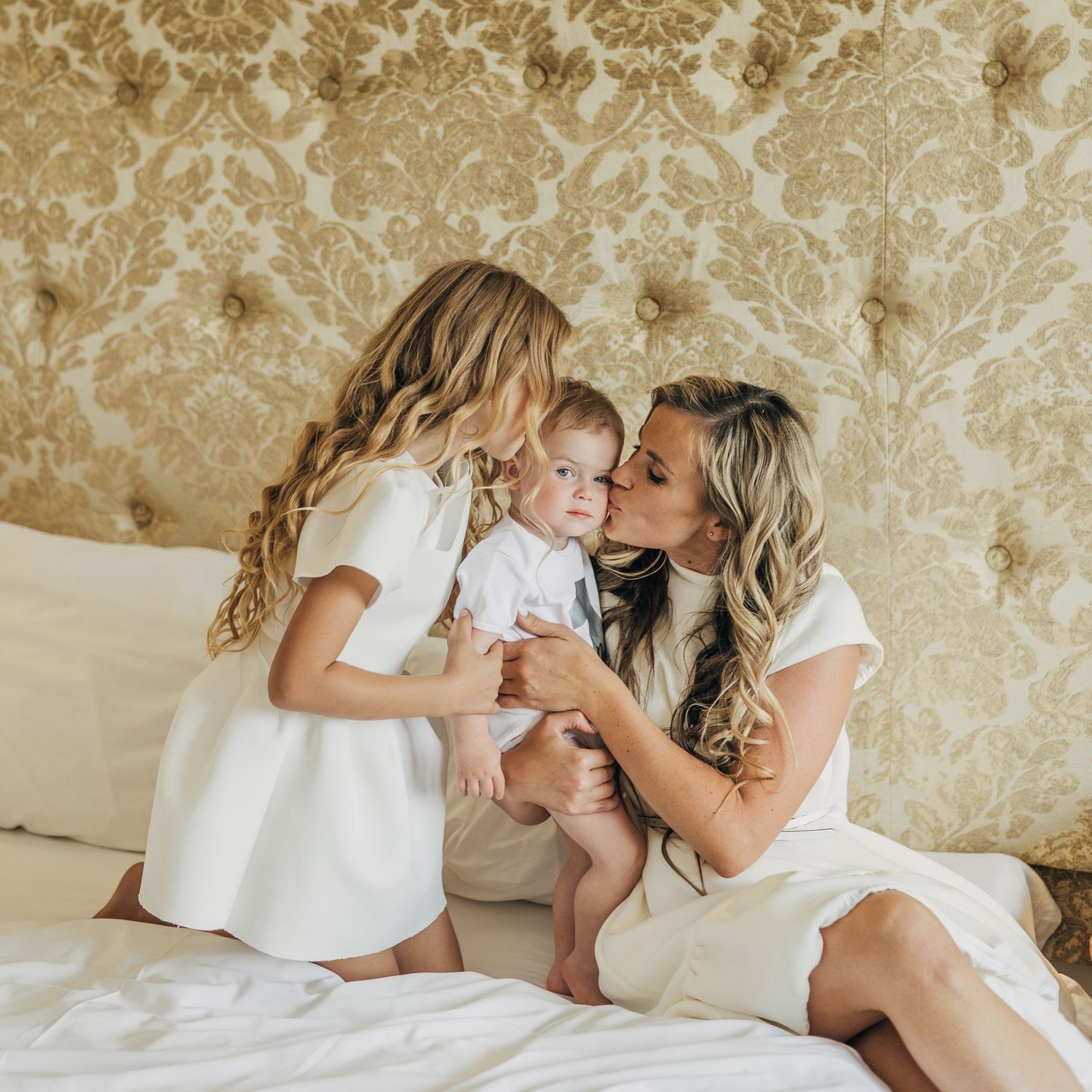 Mother kissing baby while young daughter in white dress watches on elegant hotel bed.