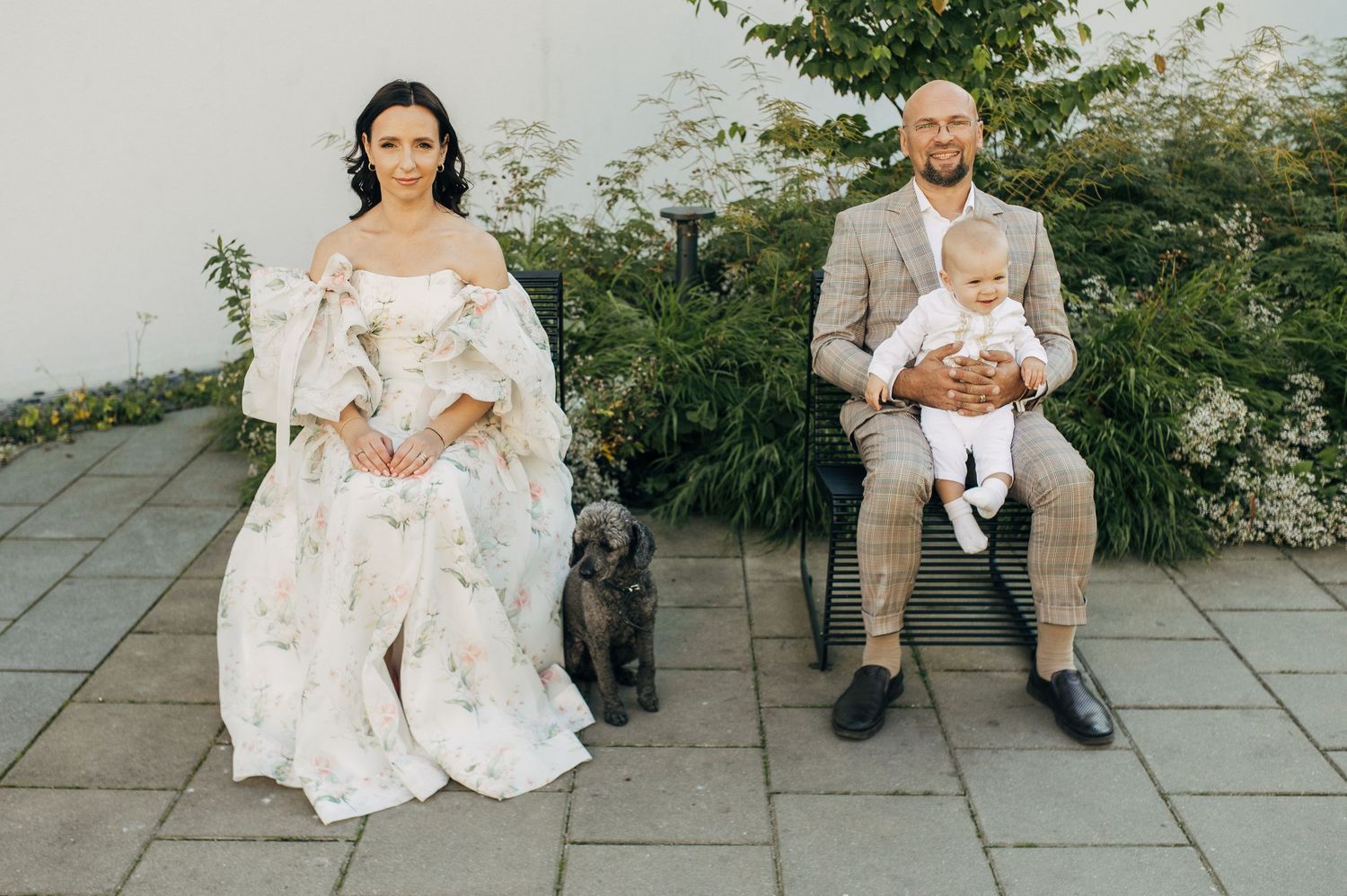 Wedding couple with baby and dog posing on patio in elegant attire.