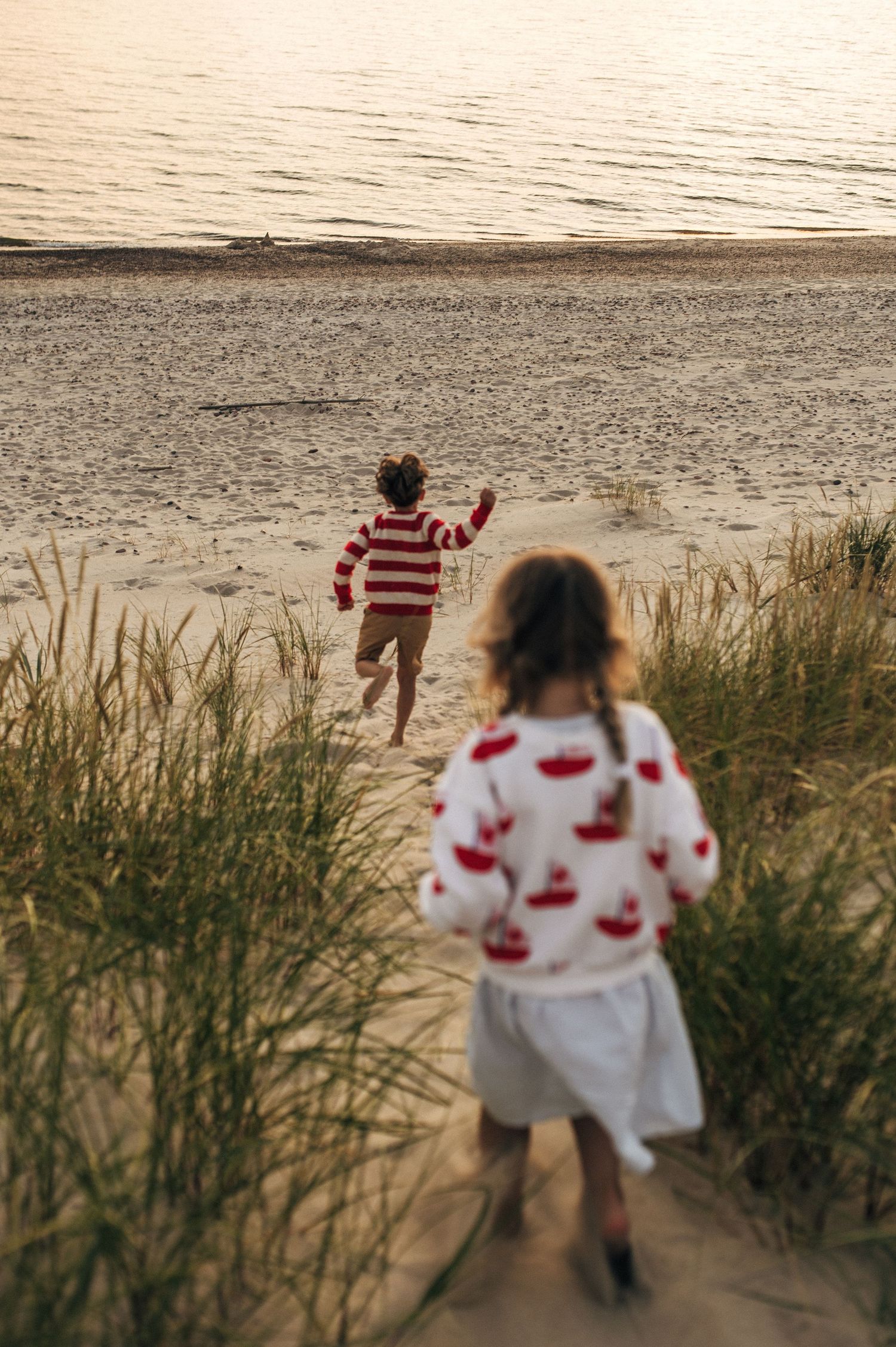 Two children running toward a sandy beach at sunset, one in red stripes, one in a floral sweater.