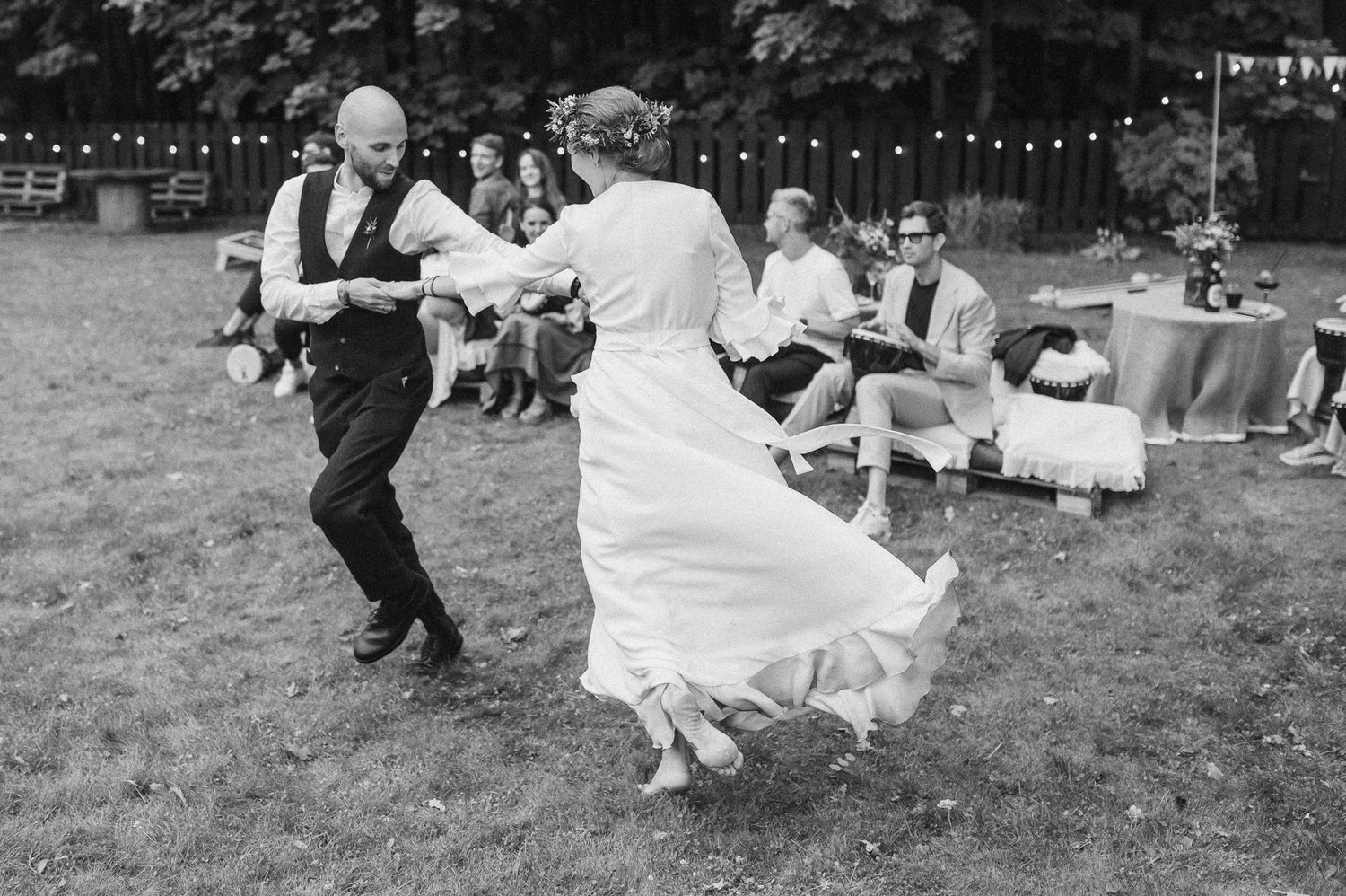 Wedding party dances joyfully on a grassy lawn with string lights overhead during an outdoor reception celebration.