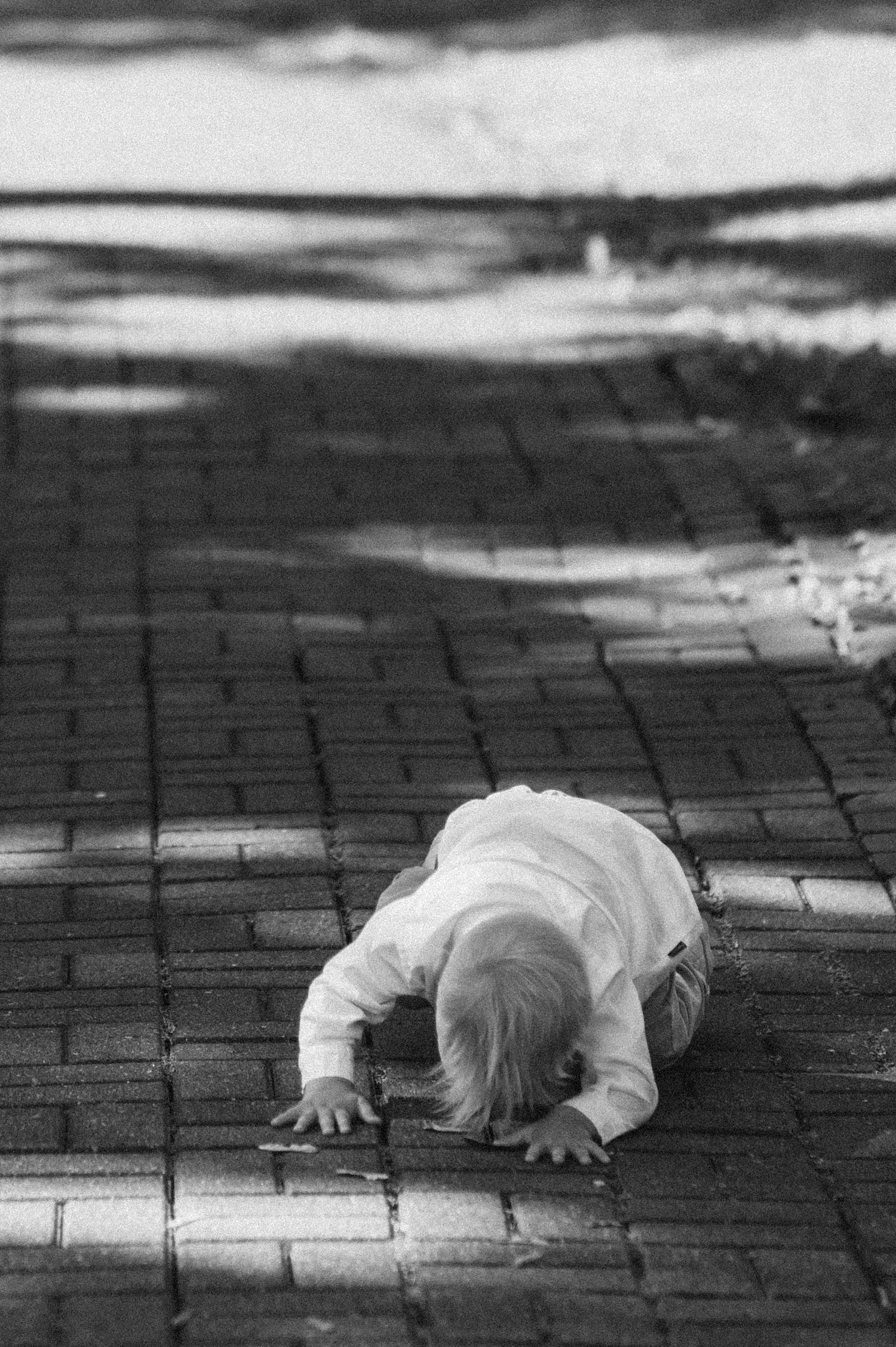 Child in white clothing crawls on brick pavement in dramatic black and white composition.
