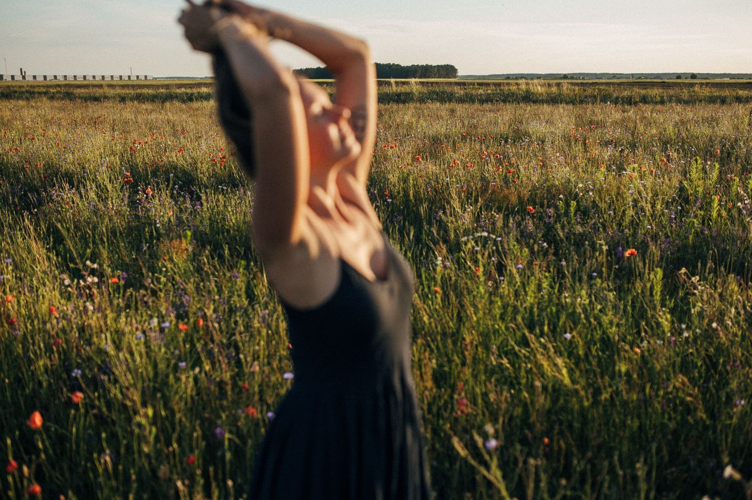 Dancing figure in sunlit meadow with arms raised against blue summer sky.