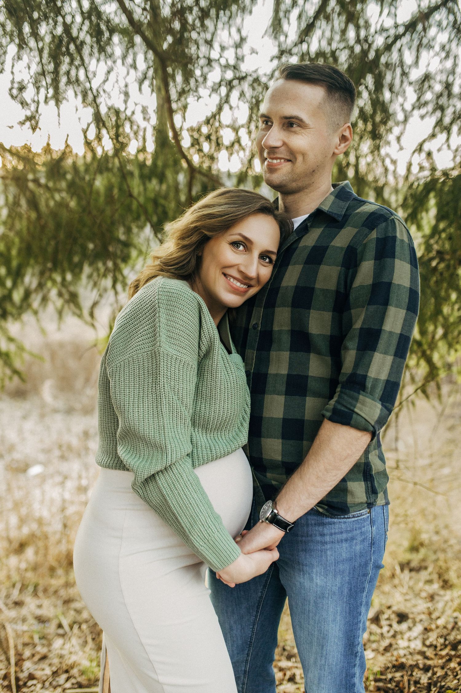 Smiling pregnant couple posing outdoors in green sweater and plaid shirt, holding hands near evergreen trees.