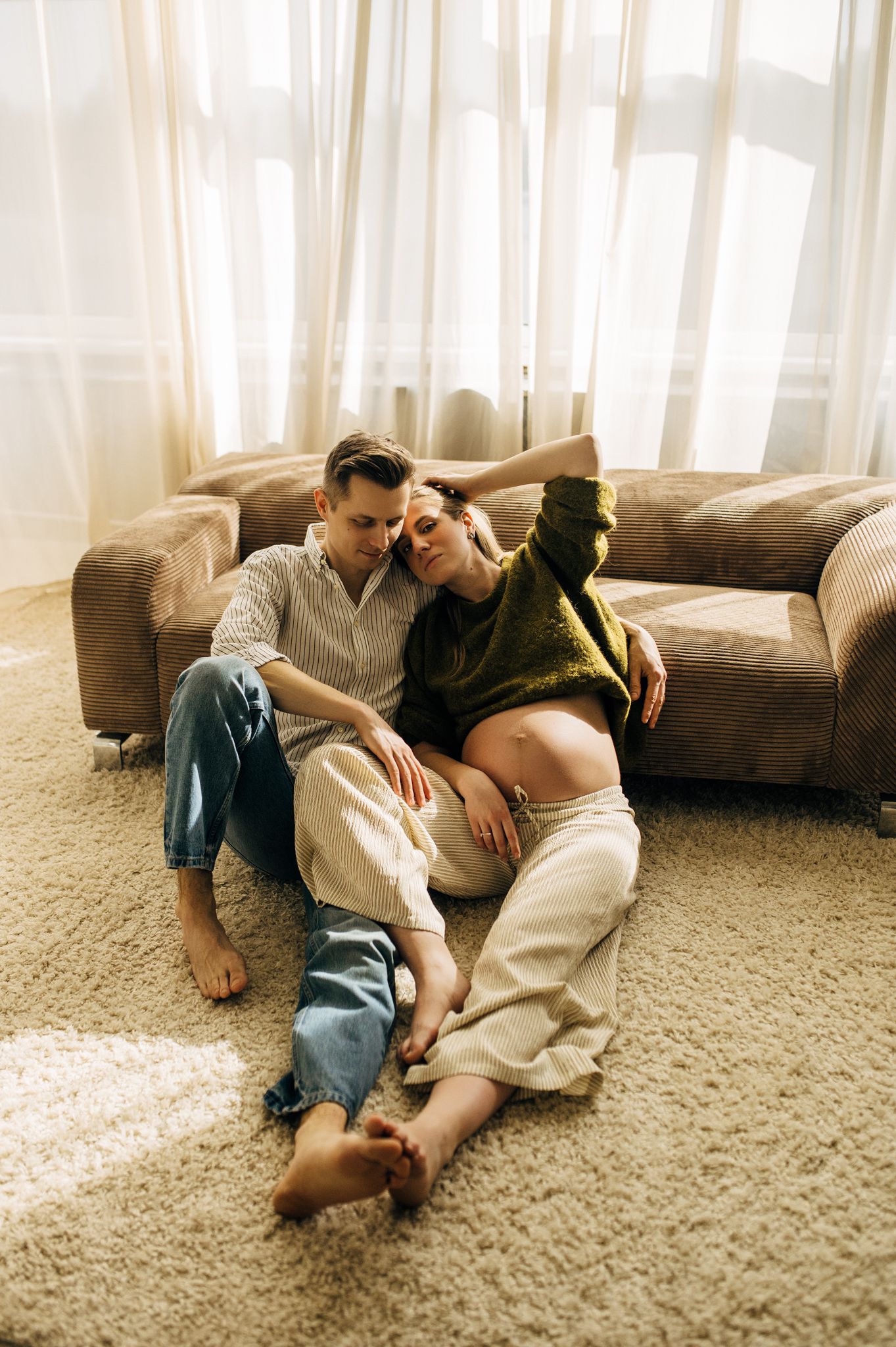 Couple sitting on carpet floor leaning against sofa, pregnant woman in green sweater relaxing with partner at home.