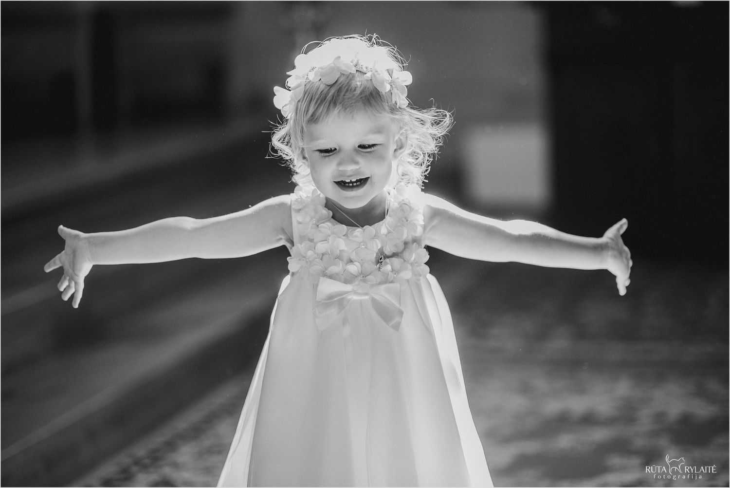 Happy toddler girl in white dress with arms outstretched, black and white photo.